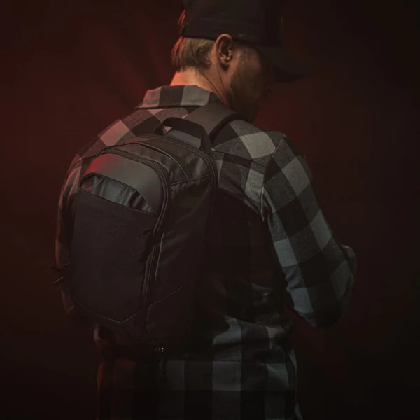 Man in a black-and-gray plaid shirt and cap, seen from behind with a backpack in dim red lighting
