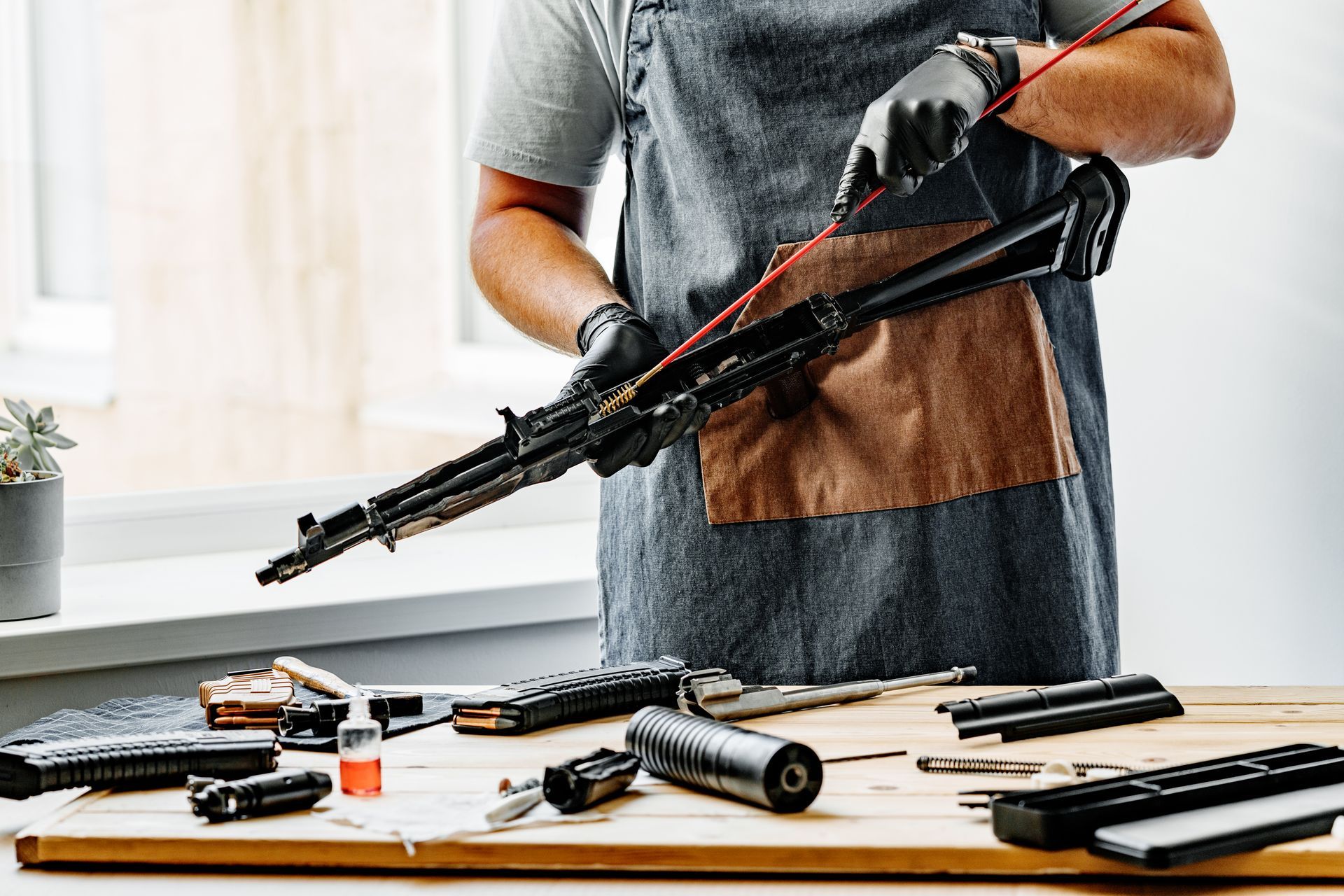 Person holding a rifle at a workbench with gun parts, magazine, and tools spread out.