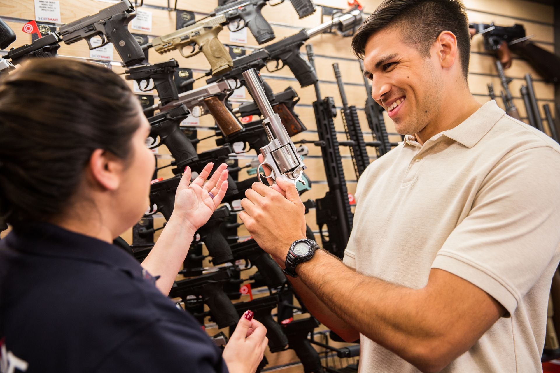 A customer examines a silver revolver while talking to a sales associate in a store filled with firearms on wall displays.