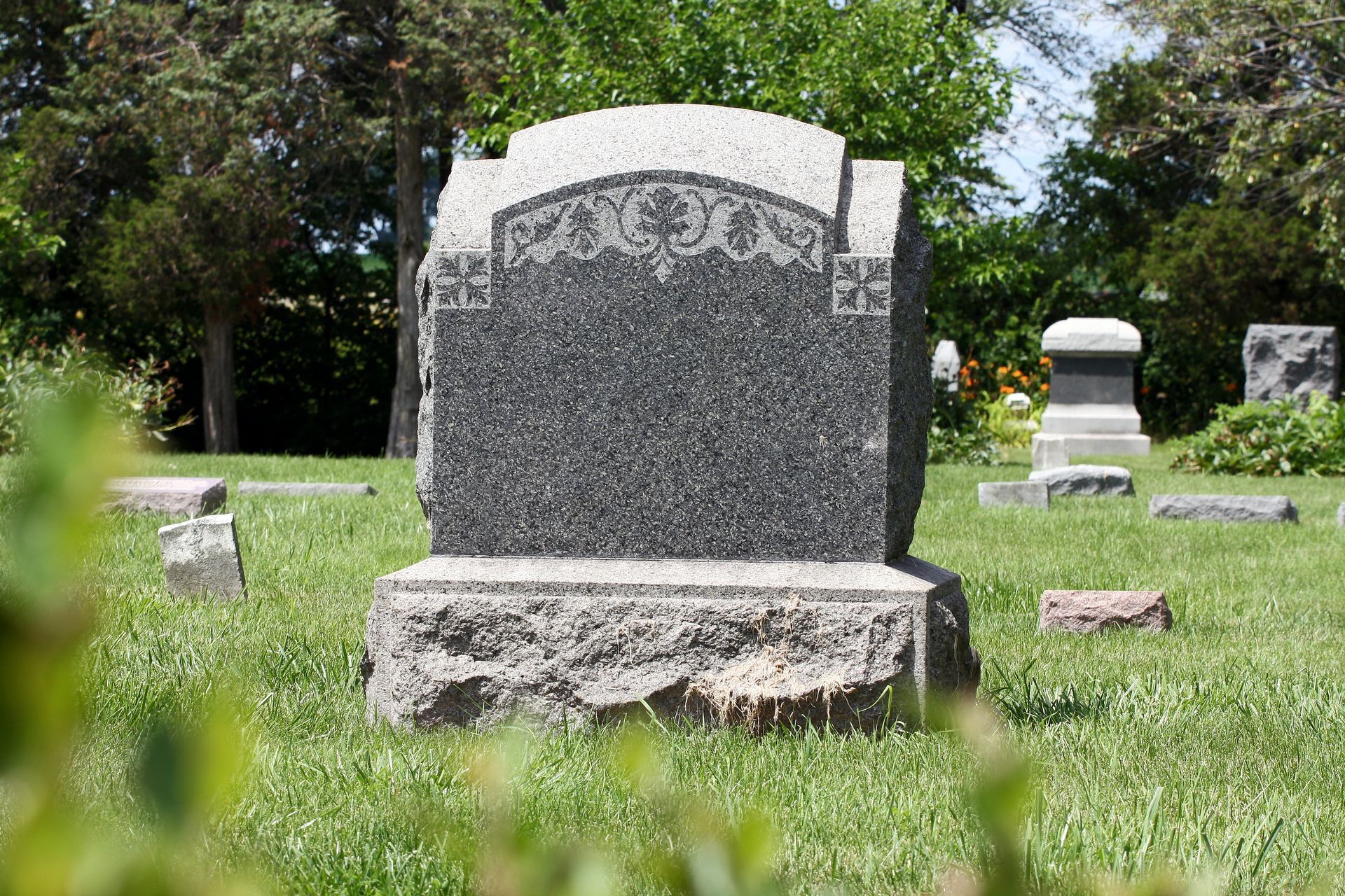 Gray tombstone in a grassy cemetery with trees and other headstones in the background Gray tombstone in a grassy cemetery with trees and other headstones in the background
