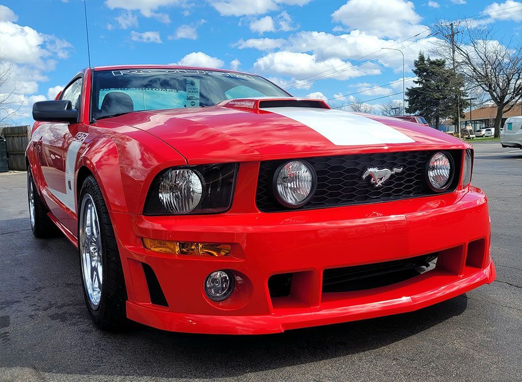 Red Ford Mustang with white racing stripes, parked on a paved surface in front of a building, under a partly cloudy sky.