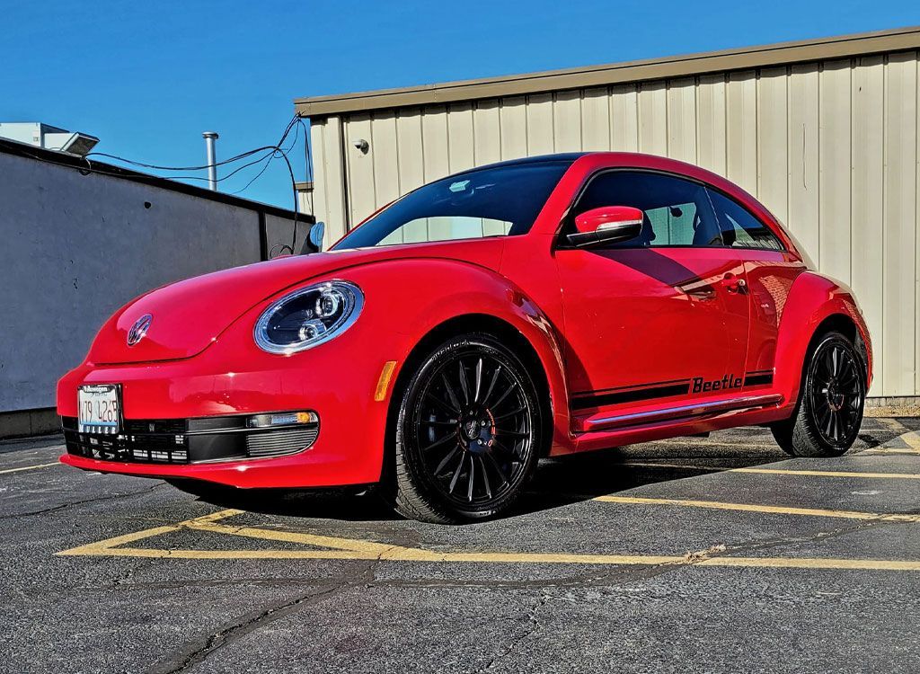 Red Volkswagen Beetle with black wheels parked in a parking lot on a sunny day. The car has a black stripe and the word