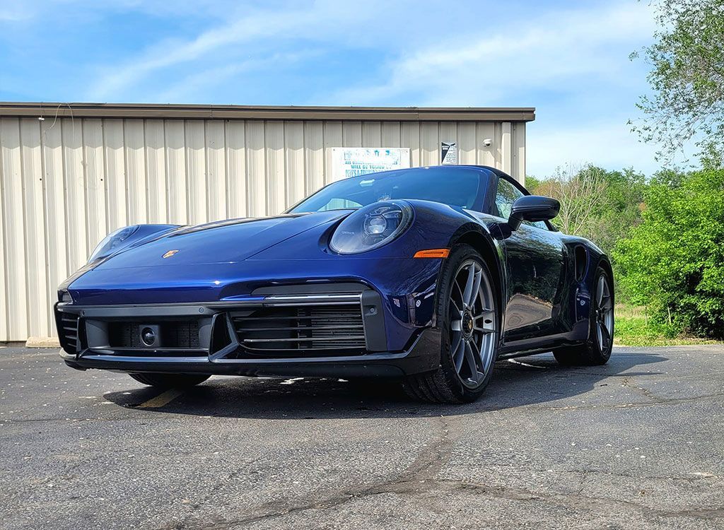 A blue Porsche 911 parked in front of a building on a gravel surface under a partly cloudy sky.