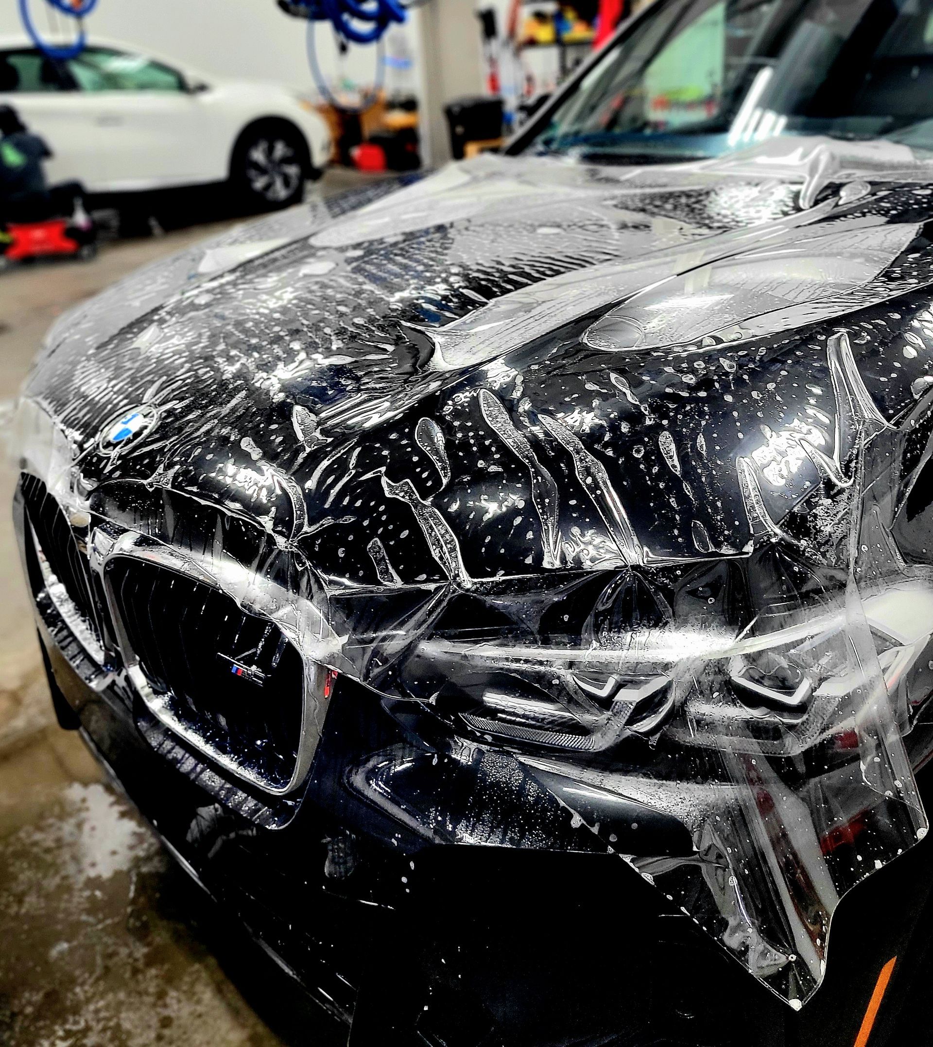 Black BMW with clear protective film being applied to the hood in a garage.