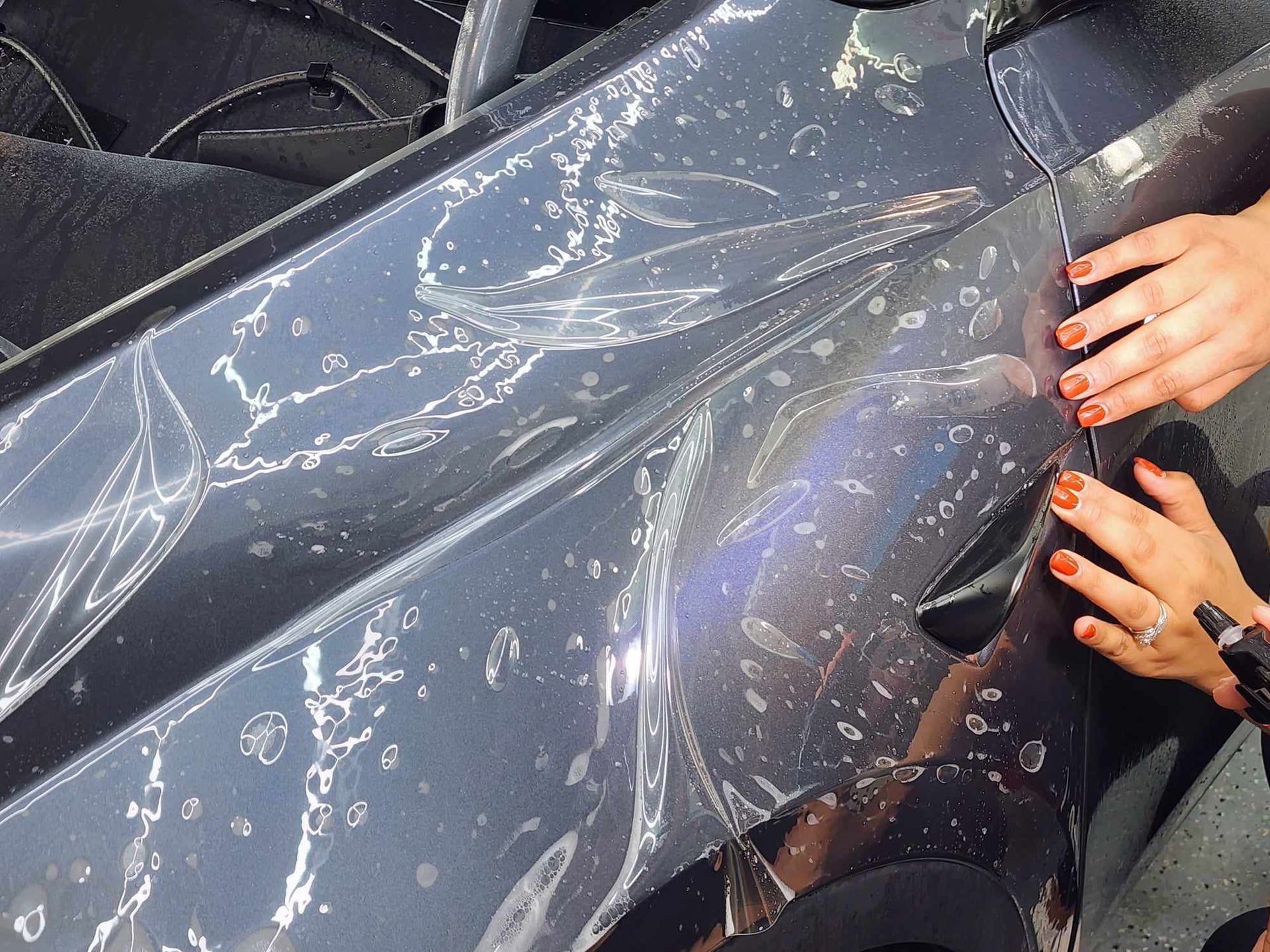 Hands applying clear paint protection film to a car's gray side panel.