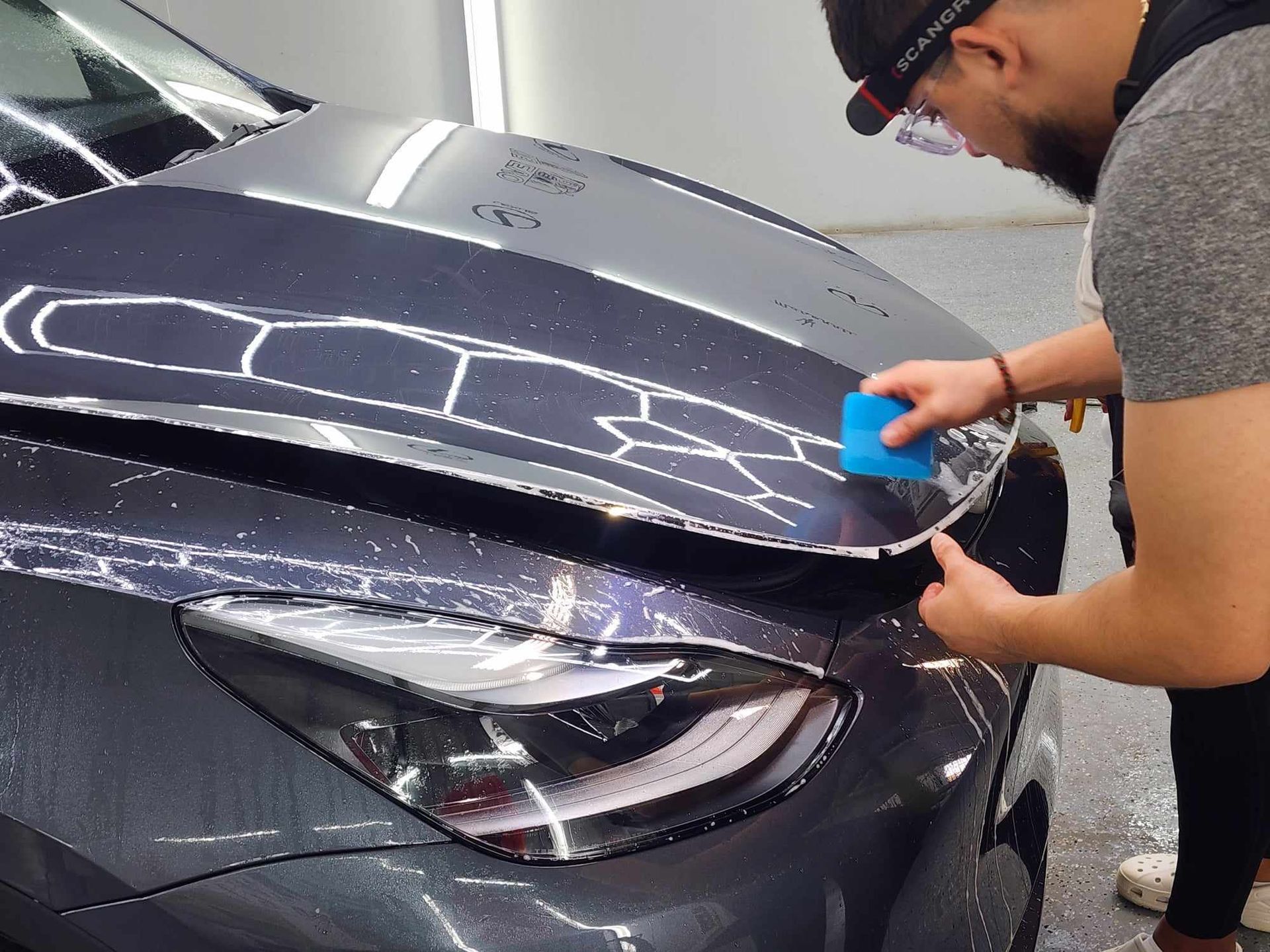 A person applies a blue squeegee to the hood of a dark gray car, likely installing a protective film.