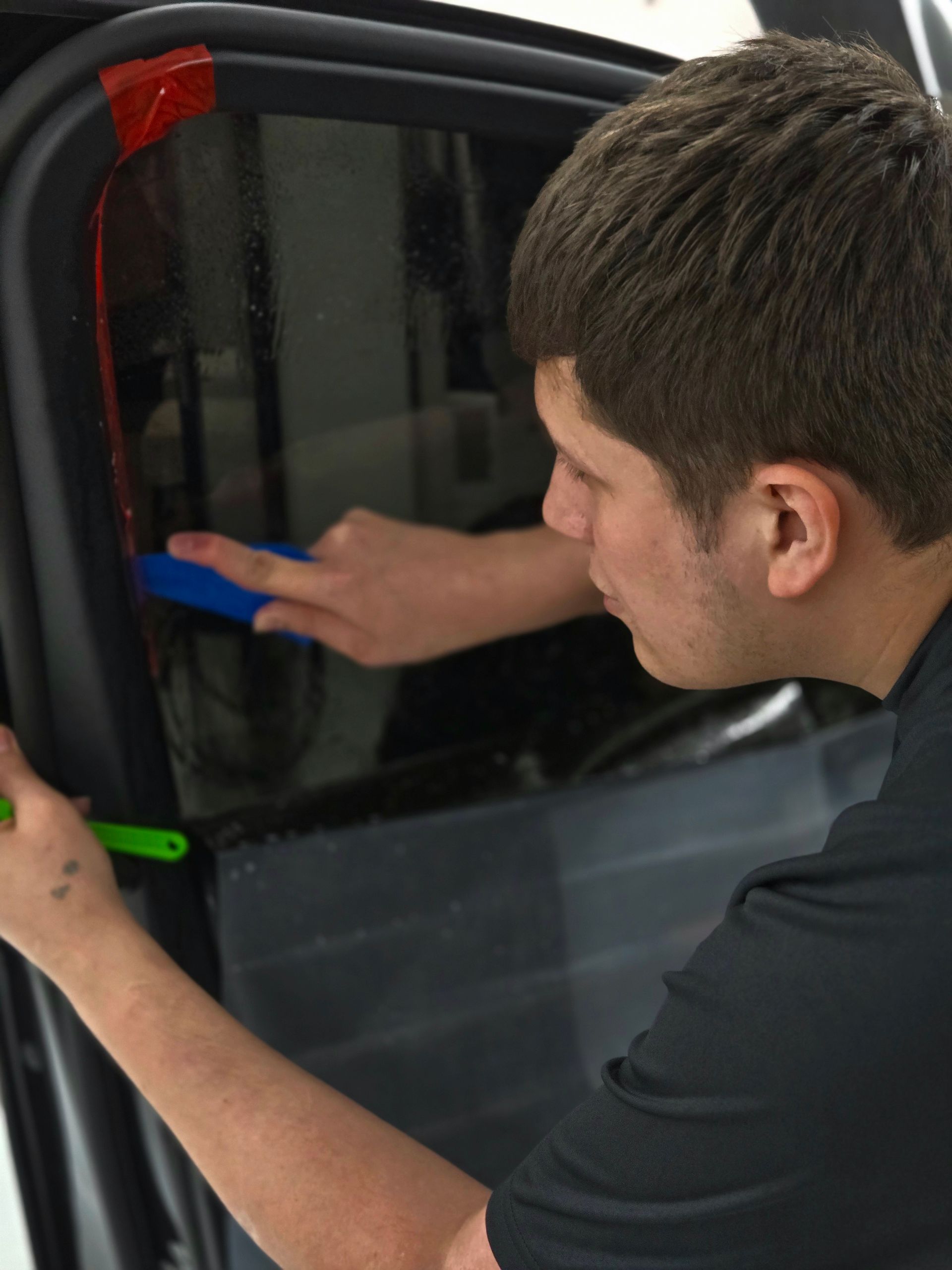 A person applying a protective film to the hood of a dark gray car in a shop.