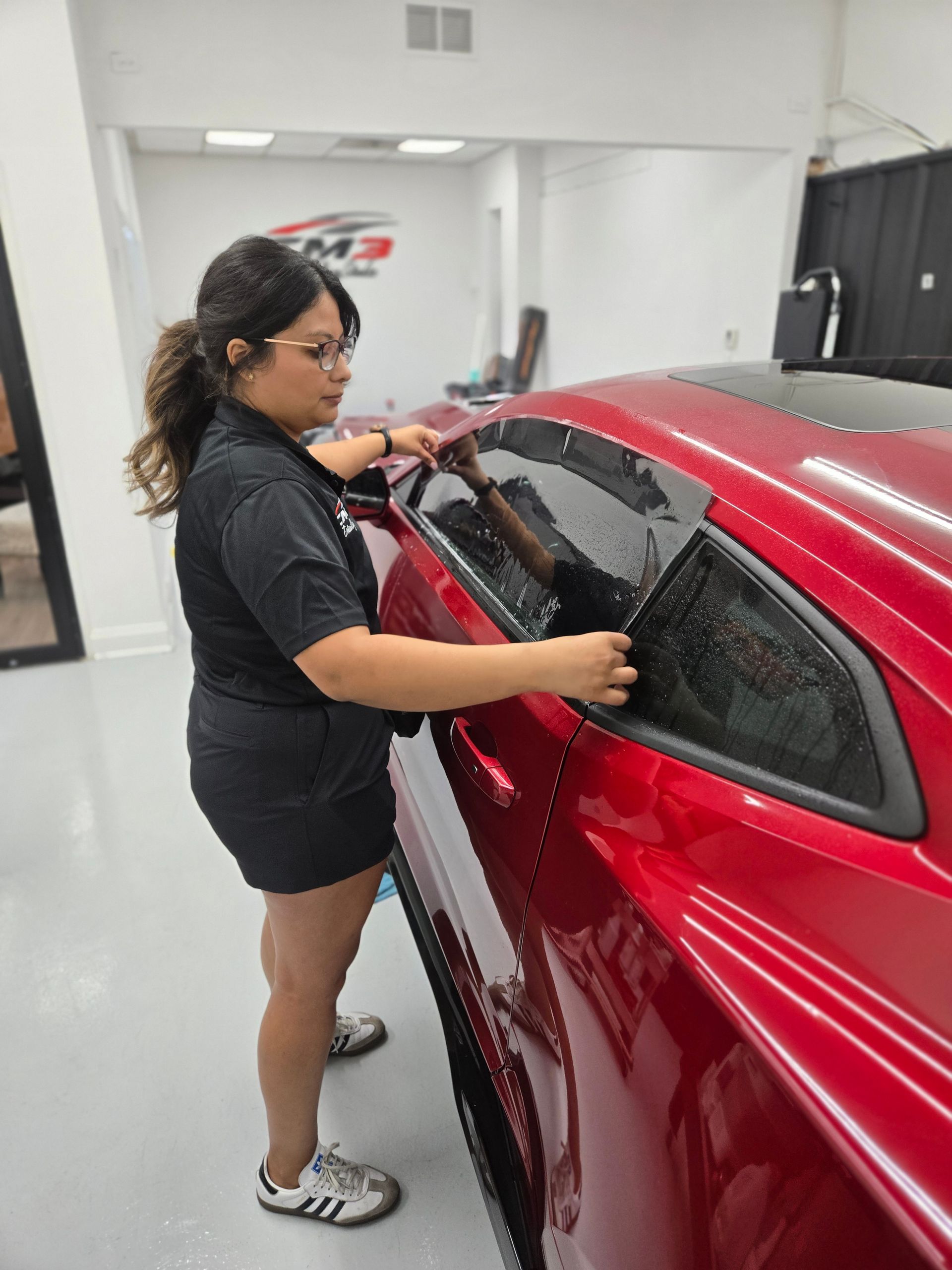 Woman applying window tint to a red sports car inside a shop.
