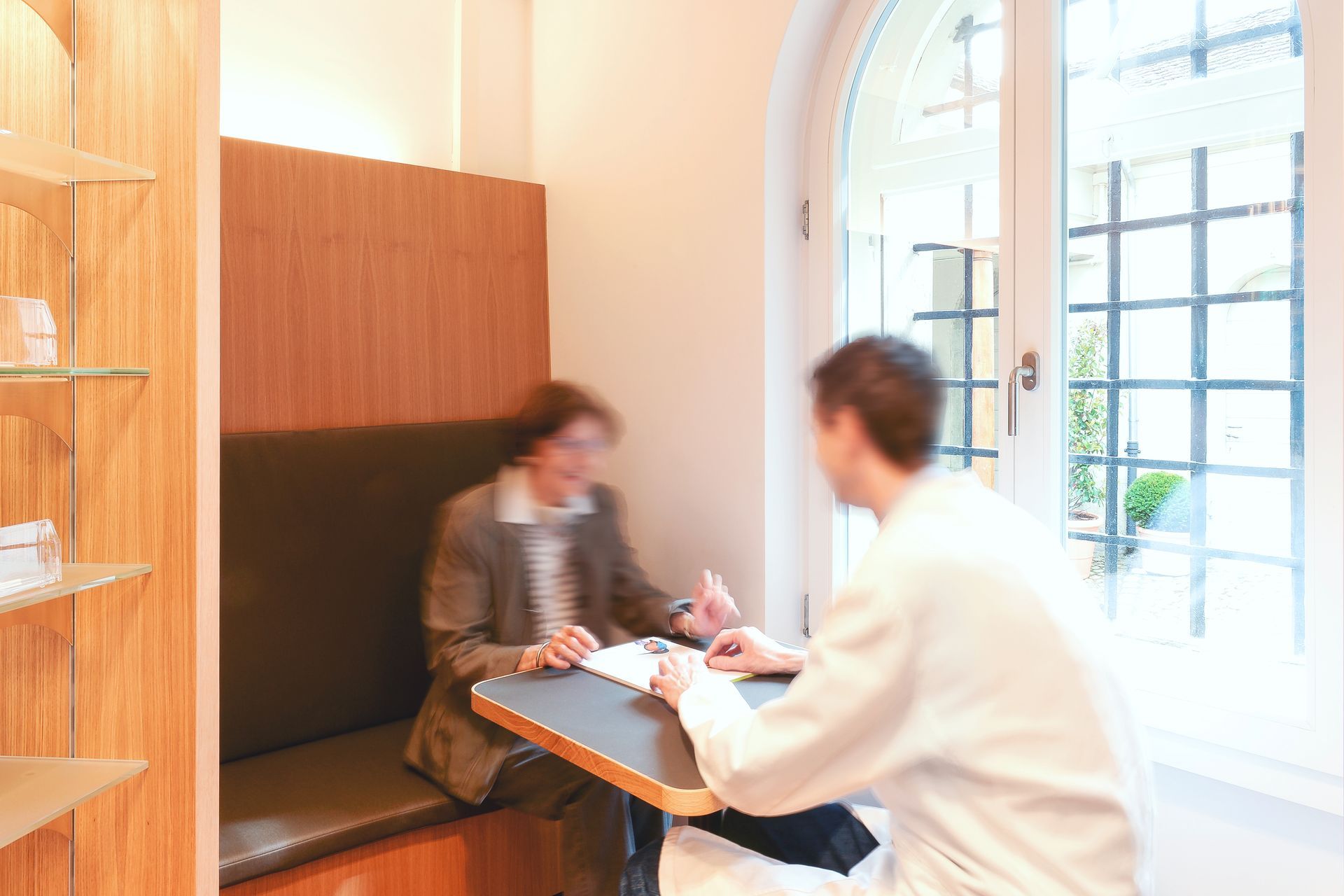 Zwei Personen treffen sich an einem kleinen Tisch in einem hellen Raum mit grossem Fenster.