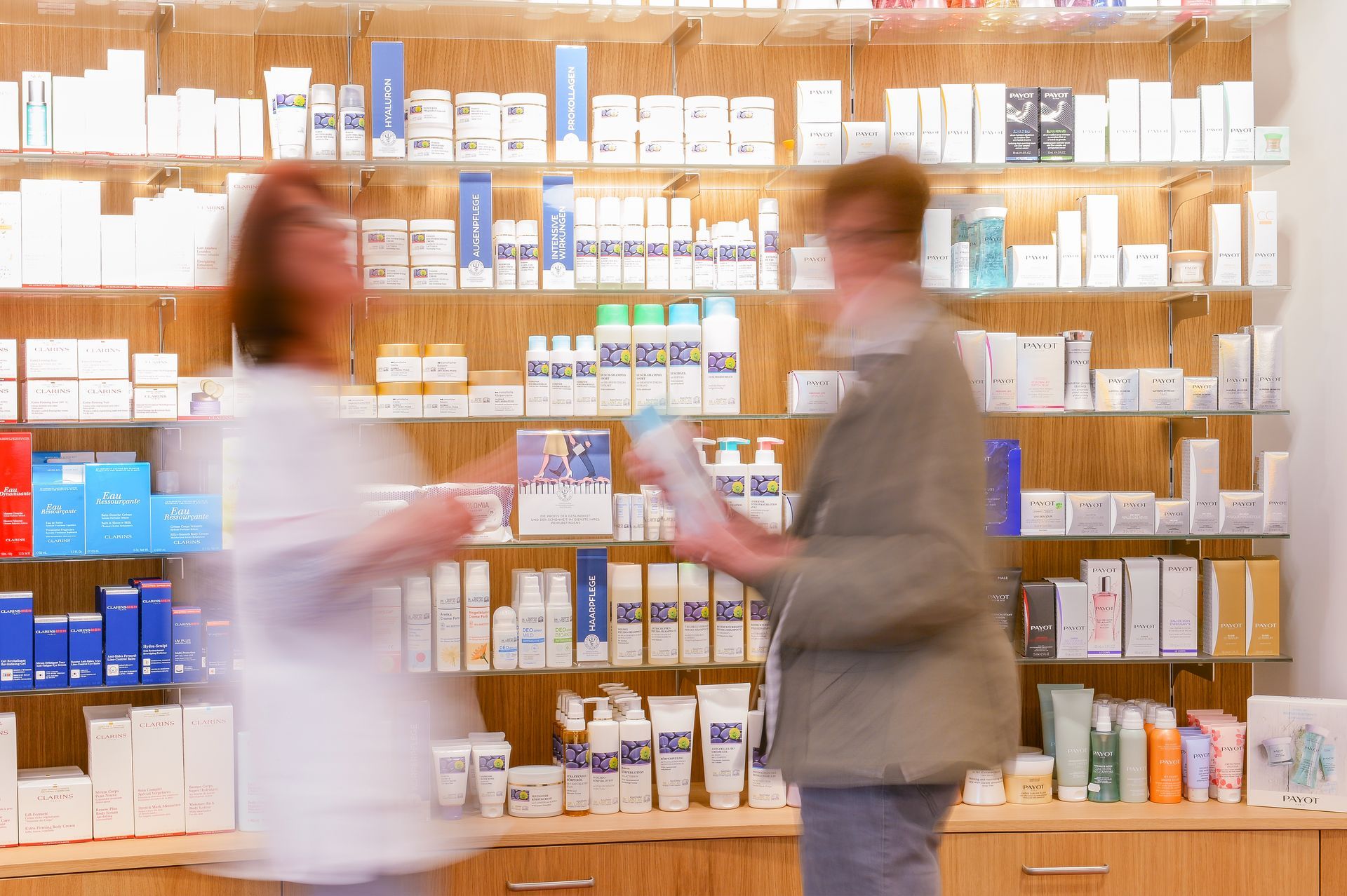 Zwei Personen unterhalten sich in einem Apothekengang, hinter ihnen stehen Regale mit verpackten Produkten.