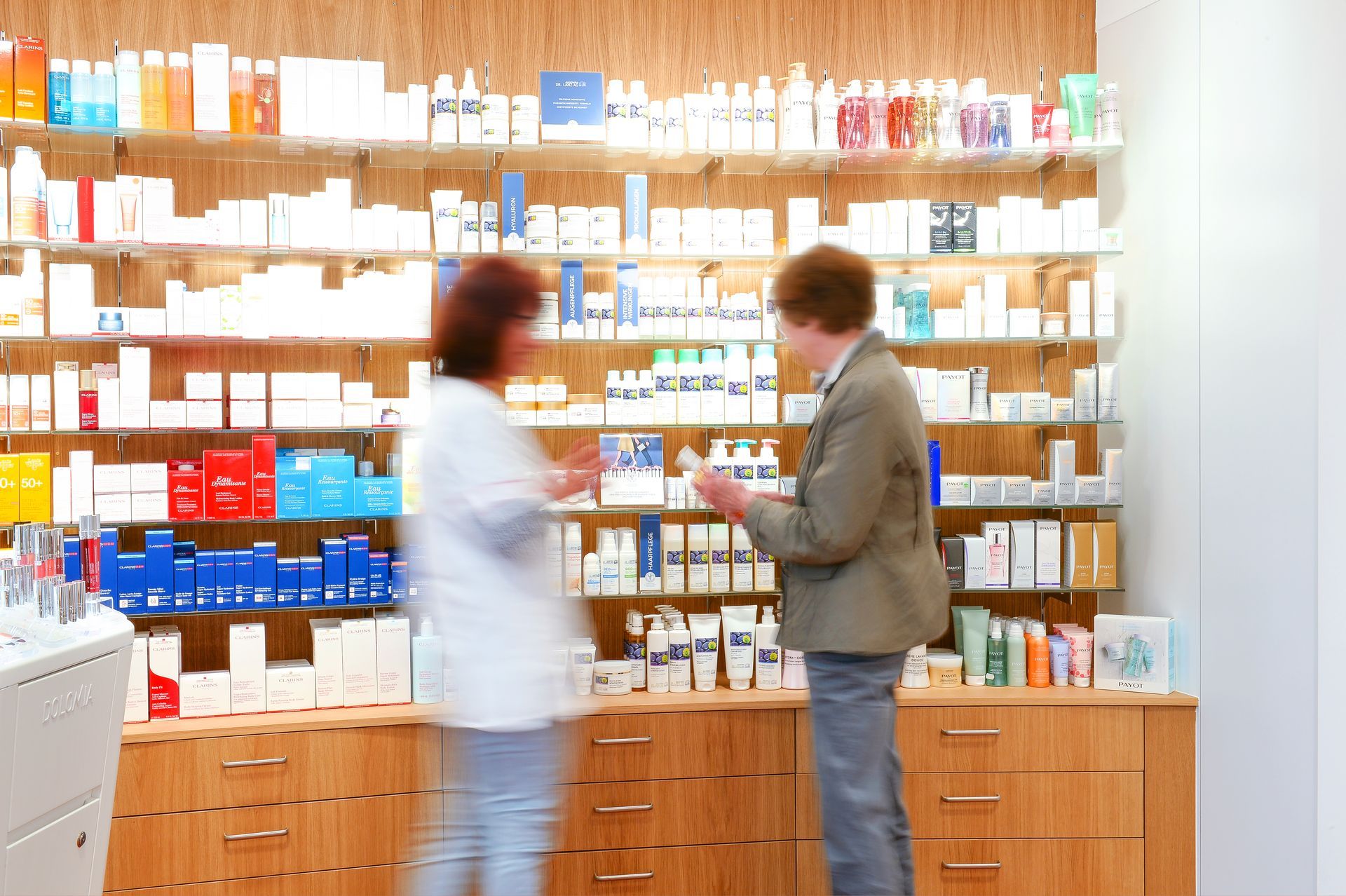 Zwei Personen unterhalten sich in einem hell erleuchteten Gang einer Apotheke, hinter ihnen stehen Regale voller Medikamente.