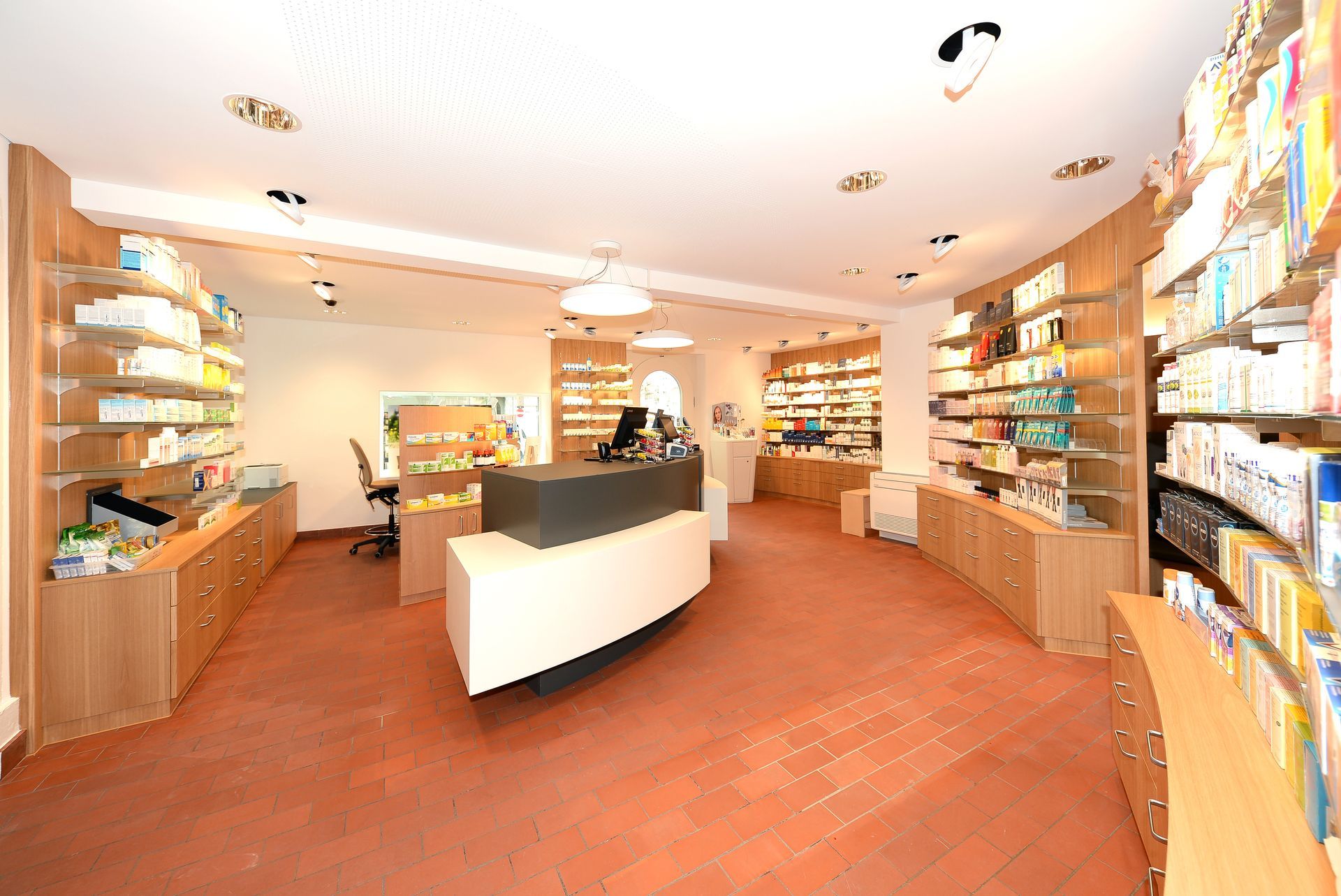 Helles Apothekeninterieur mit Holzregalen, weissem Tresen und im gesamten Laden ausgestellten Produkten.