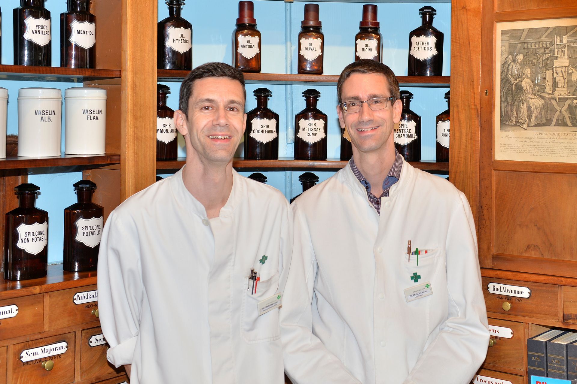 Zwei Apotheker in weissen Kitteln stehen in einer Apotheke, hinter ihnen Regale mit Medikamentenflaschen.