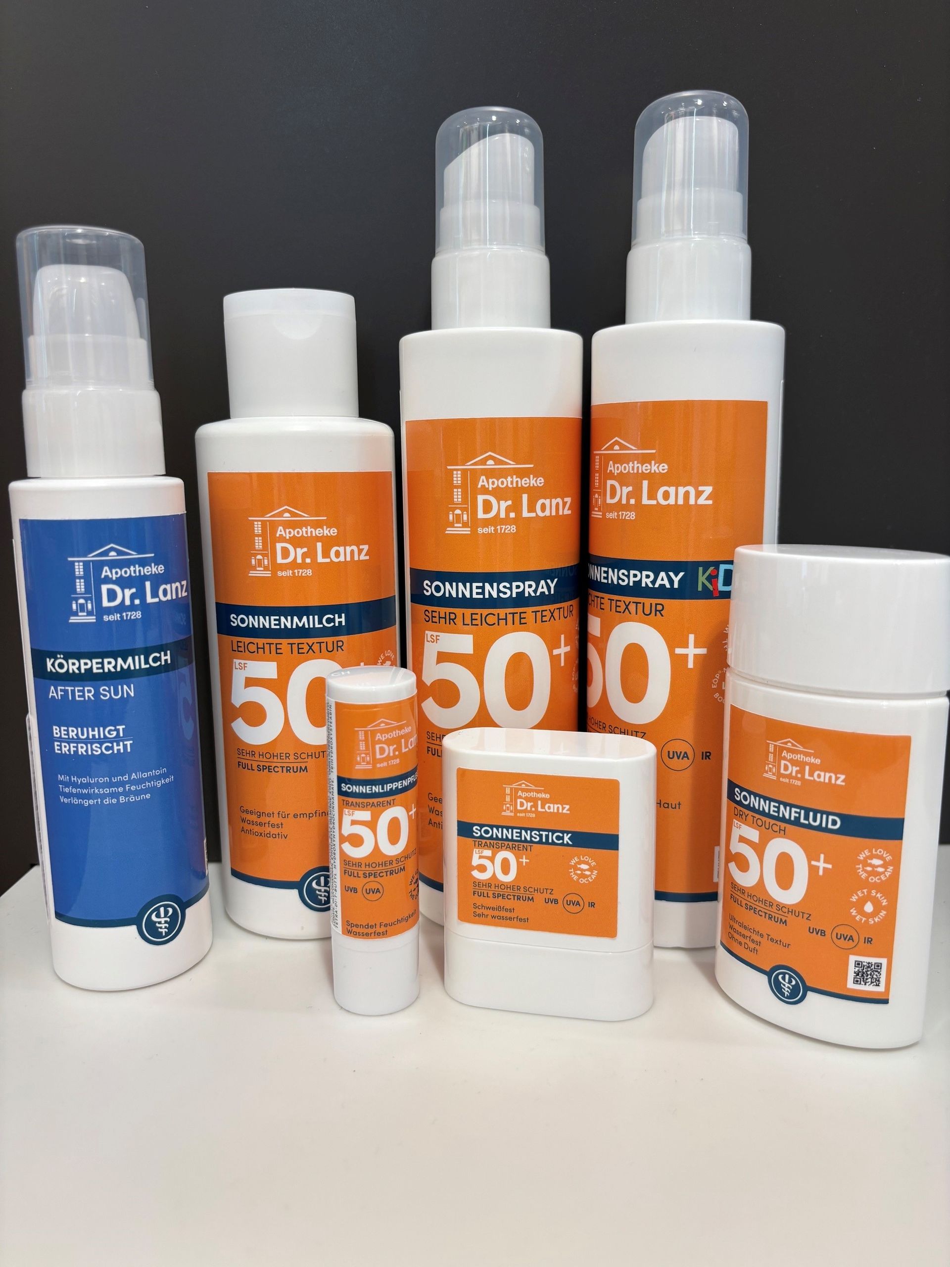 Verschiedene orange und blaue Sonnenschutzmittelflaschen und -tubes sind auf einer weissen Fläche ausgestellt.
