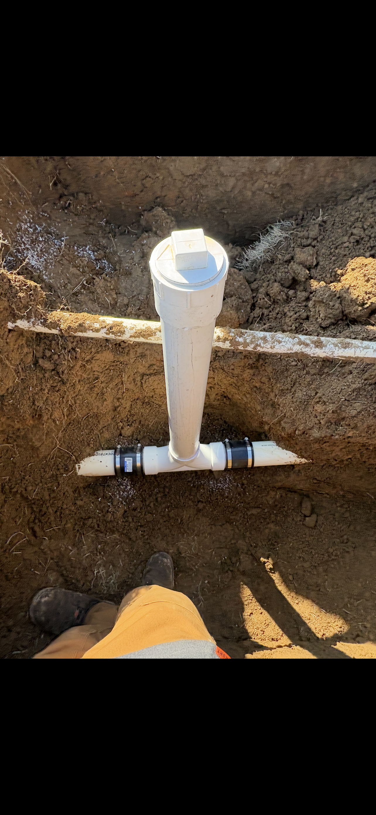 White PVC pipe T-fitting in a trench, connecting to other buried pipes.