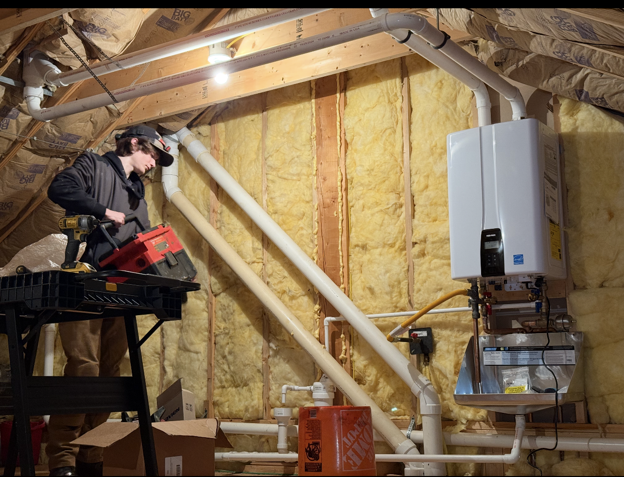 Man installing a wall-mounted water heater in an attic, working with tools. PVC pipes and insulation visible.