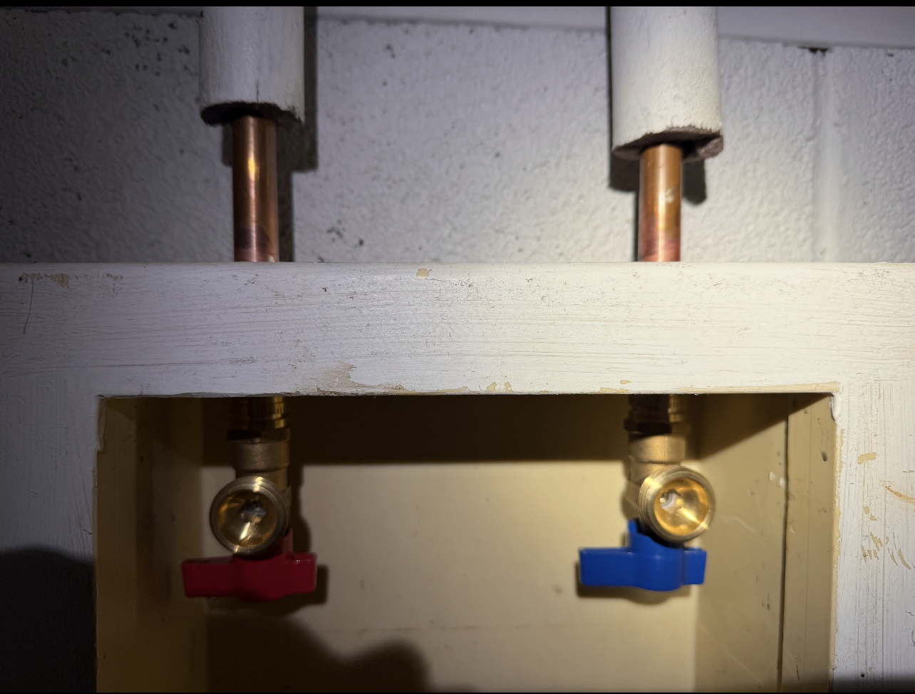 Copper pipes with red and blue shut-off valves for hot/cold water in a wall box.
