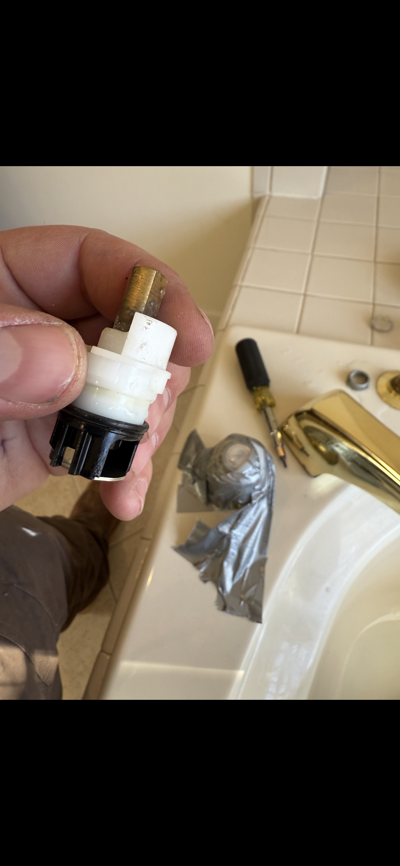 A hand holding a white and black faucet valve cartridge. A faucet is on the right. Bathroom setting.