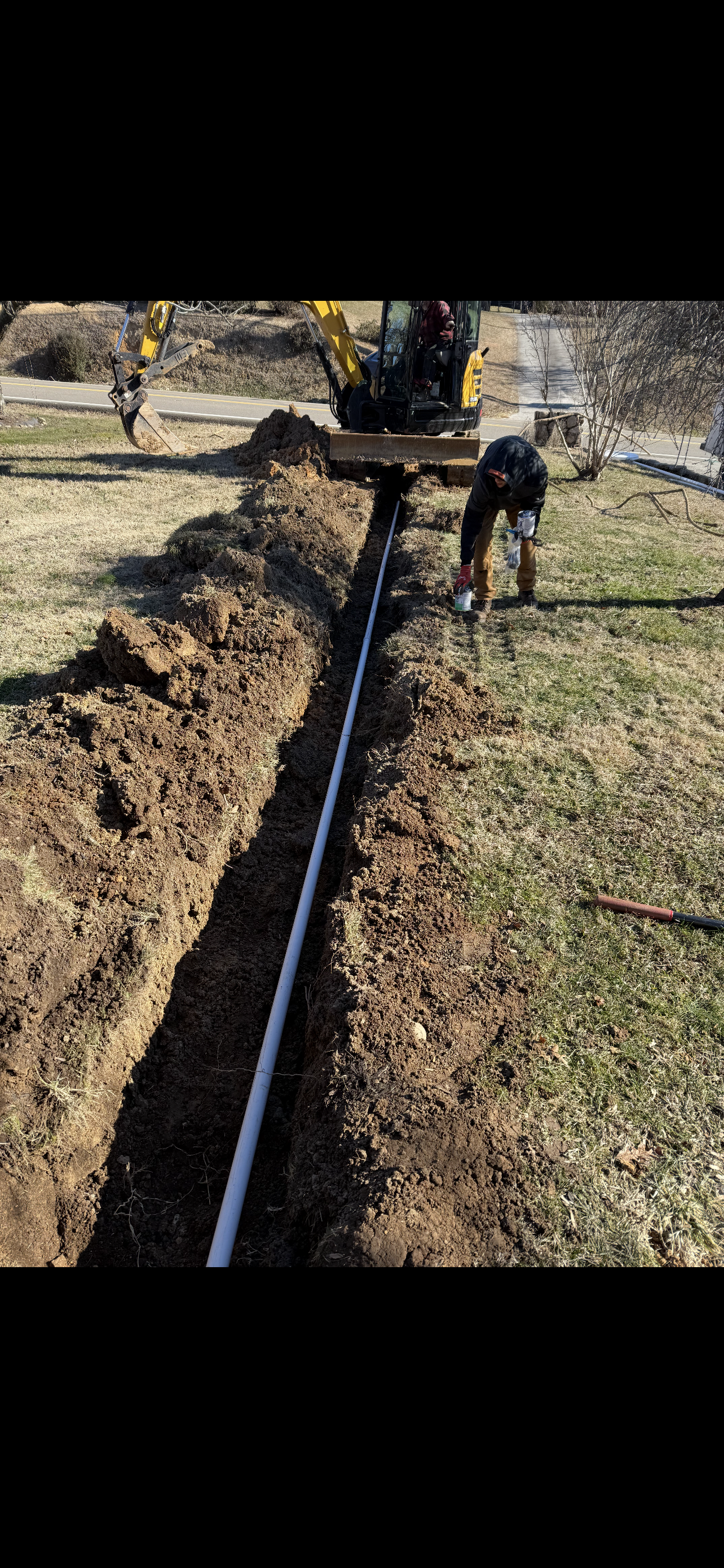 A trench in the ground with a pipe being installed, a small excavator and a worker present.