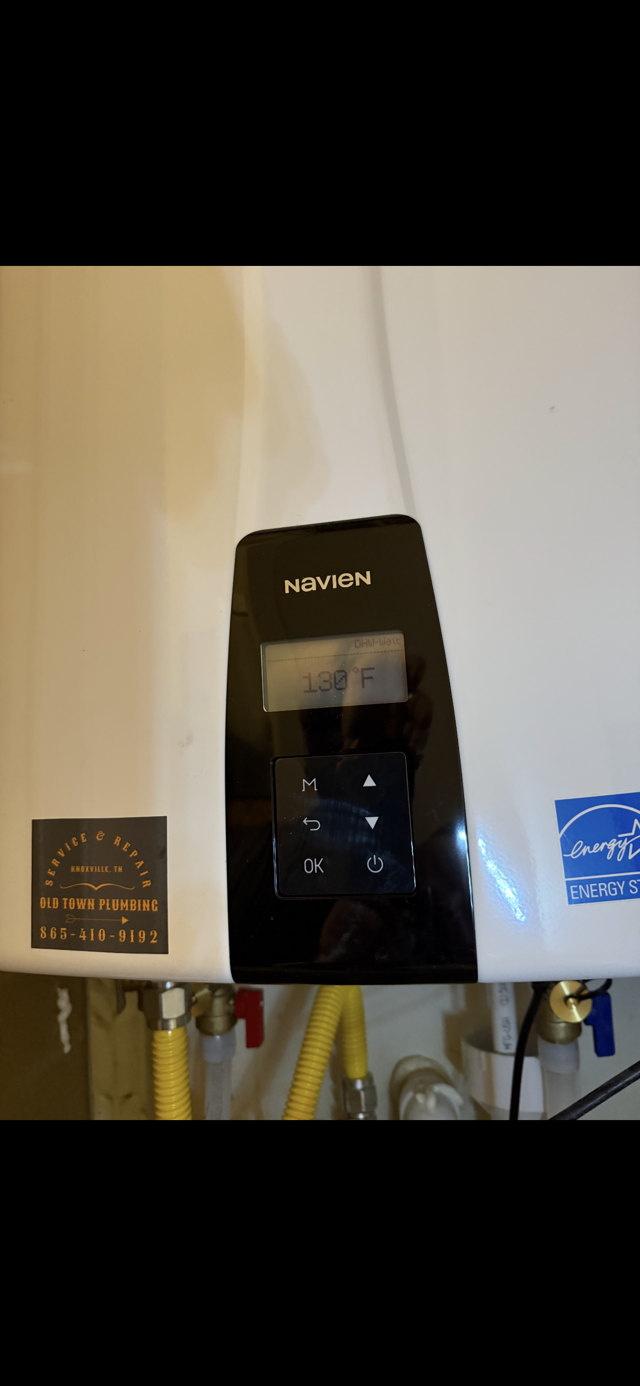 A white water heater with a black control panel, pipes, and labels are visible.