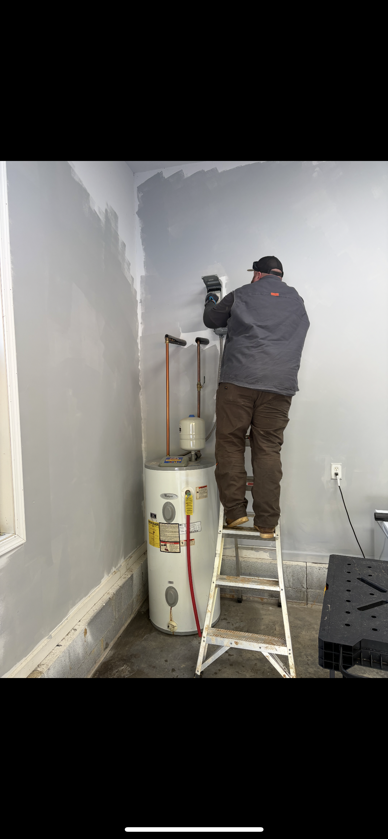 Person on ladder working on pipes above a water heater in a corner, indoors. Gray walls, concrete floor.