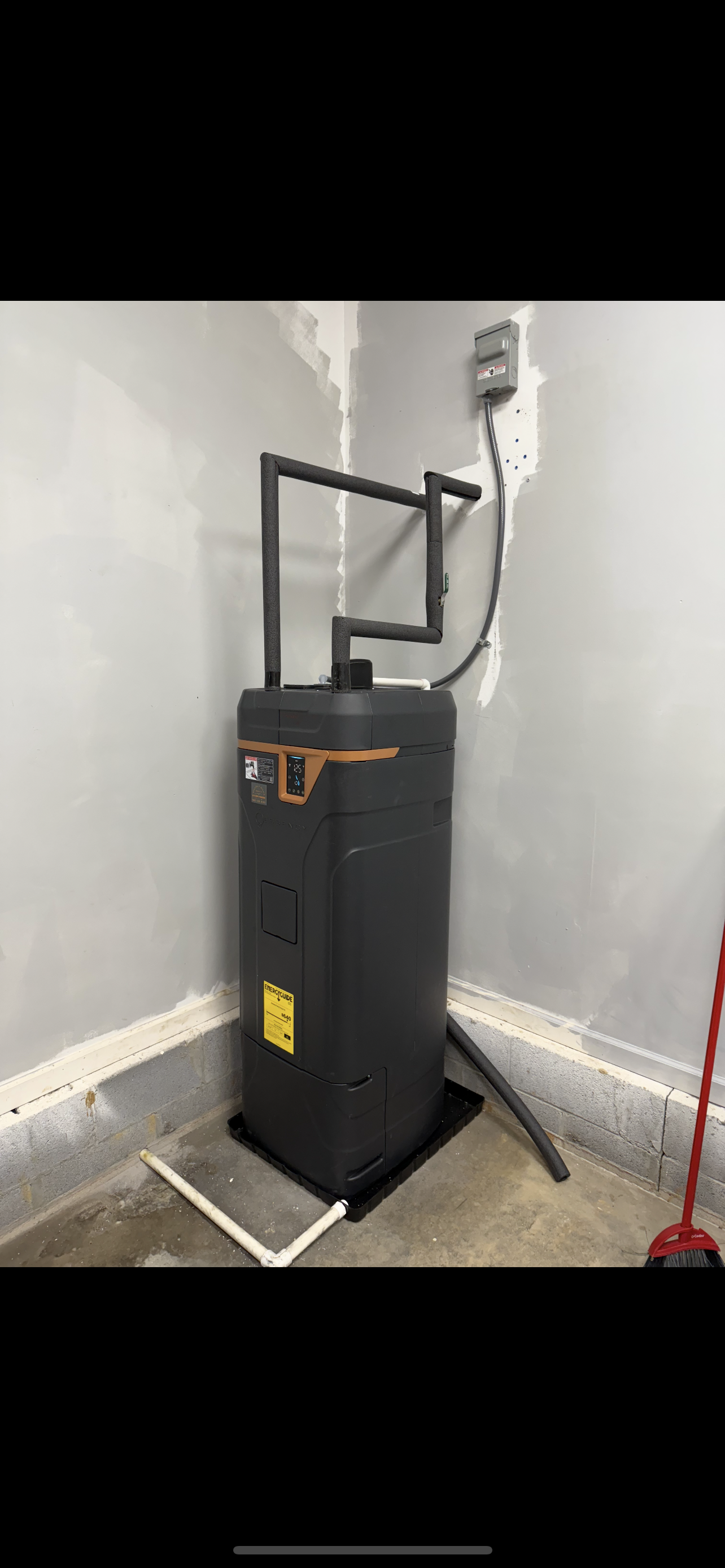 Black water heater in a corner of a room, near a wall outlet, with a pipe and broom.
