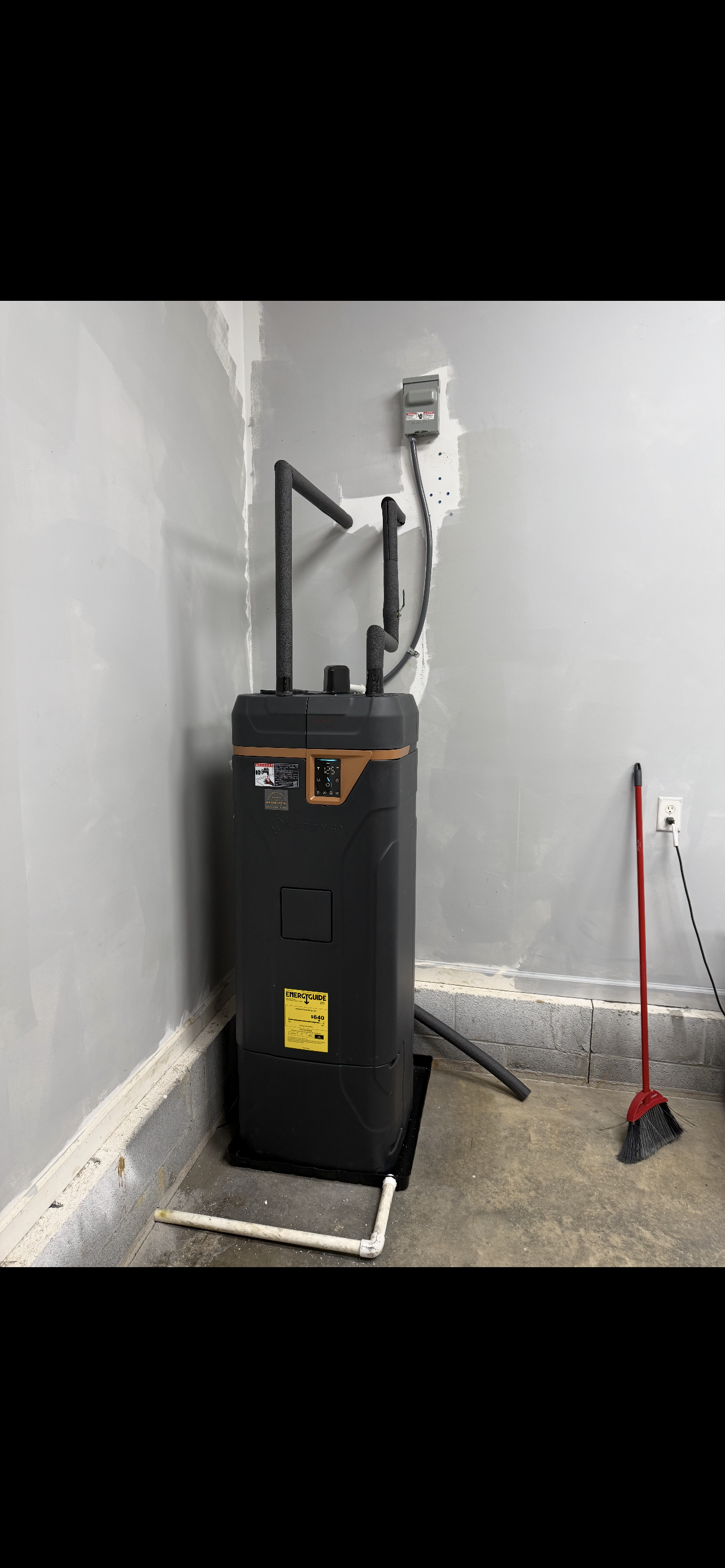 A black water heater in a corner of a room with a broom and piping.