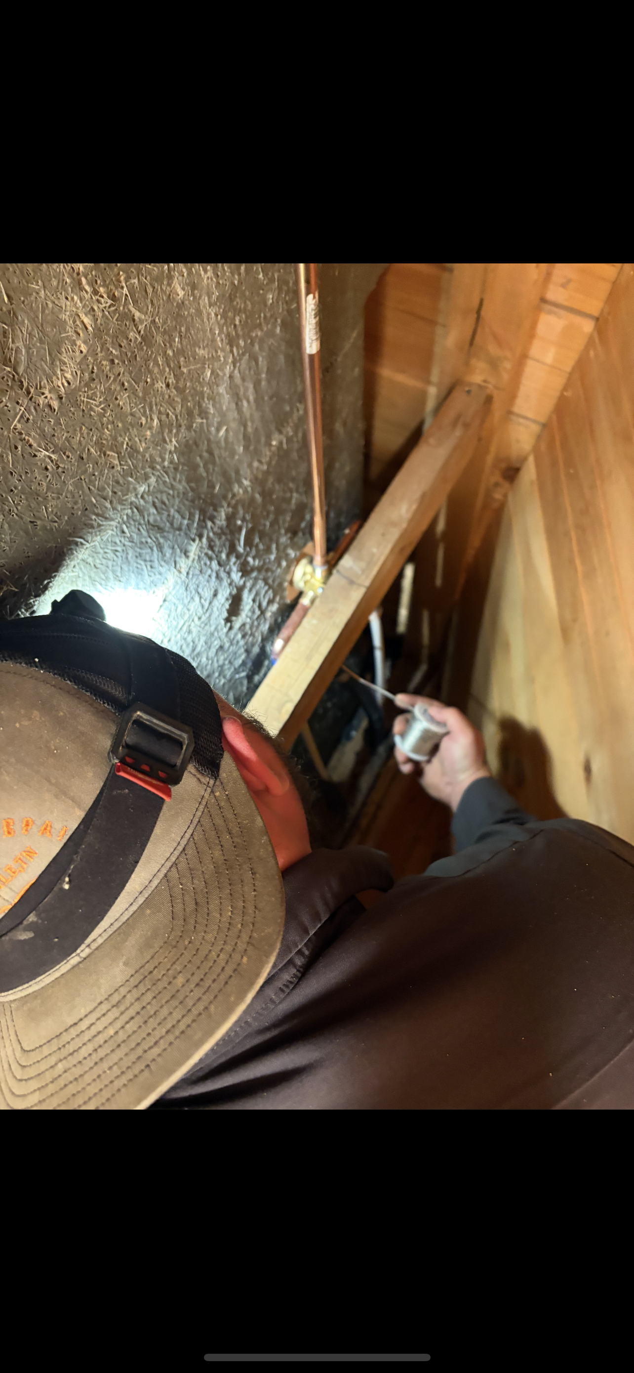 Person working on wiring in a dark attic. He wears a hat, and is holding a tool.
