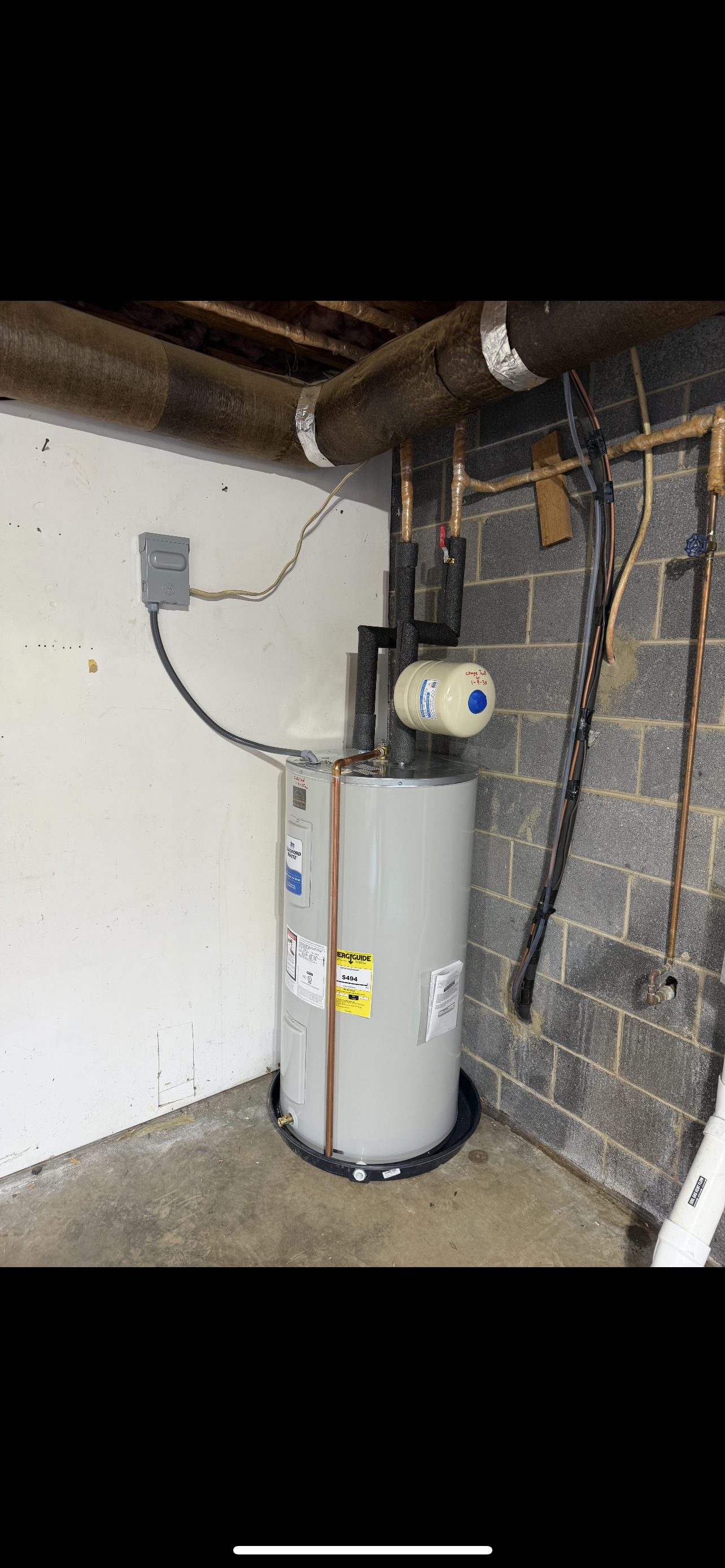A tall, cylindrical water heater in a basement corner, with various pipes and wires.