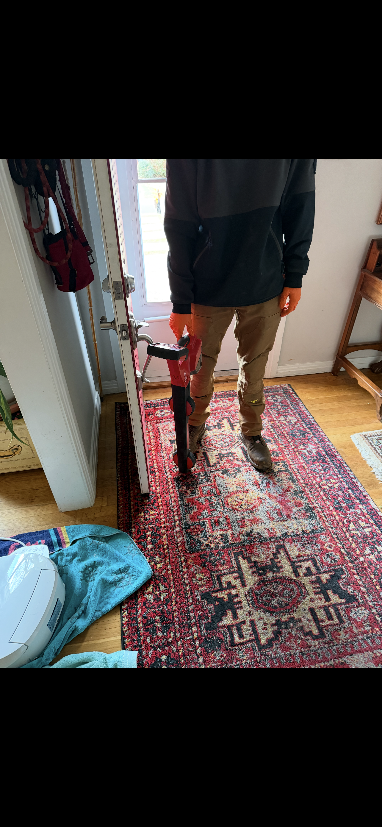 A person standing on a red rug near a window, wearing a black and grey hoodie, and brown pants.