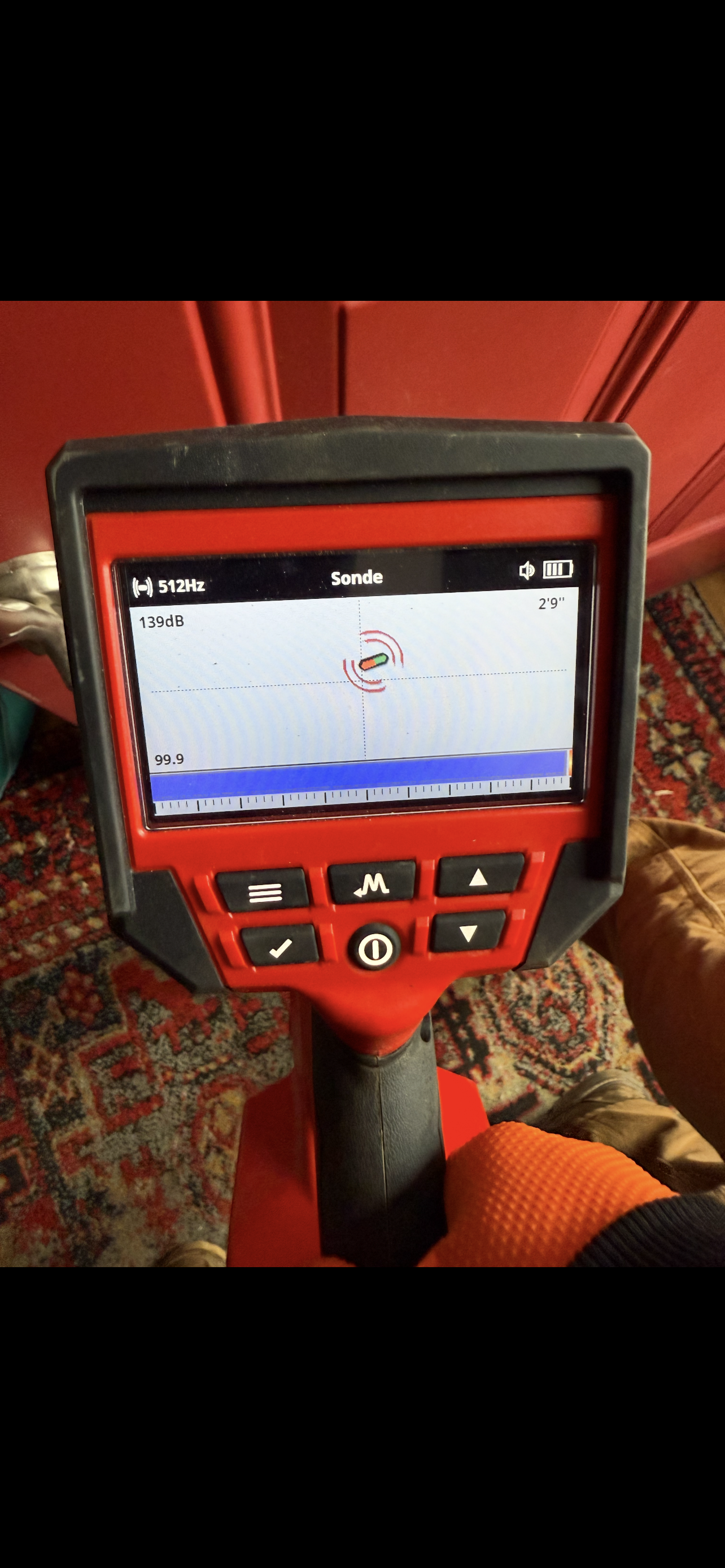 A person holding a red and black device with a screen showing sonar readings, likely a stud finder.
