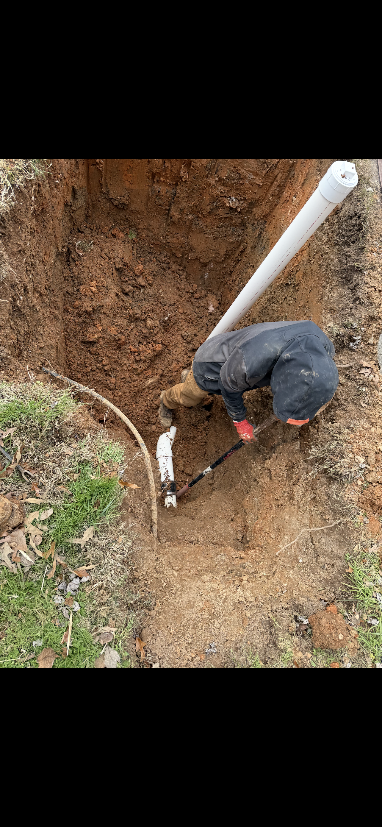 Person digging in a trench, working on a pipe outdoors.