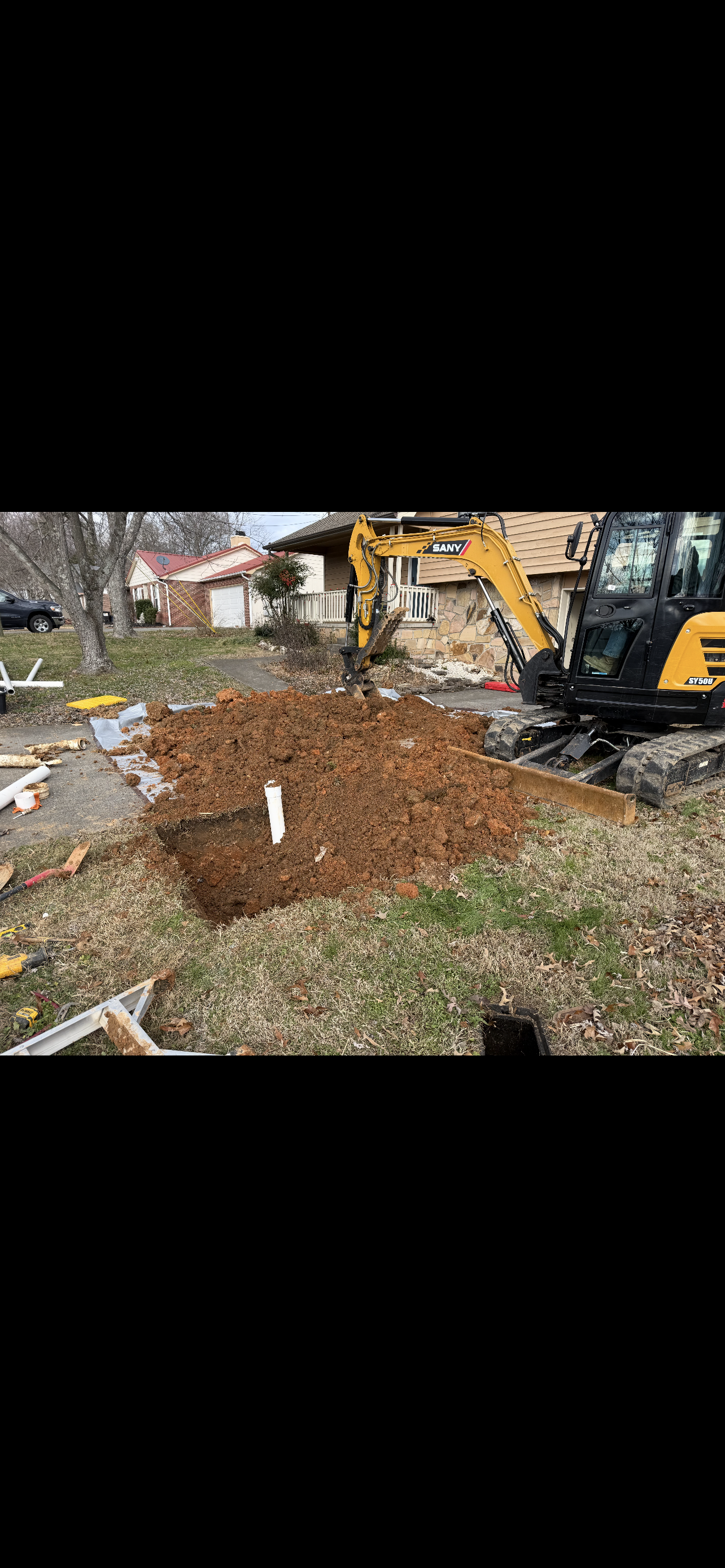 An excavator digs in a residential yard, near a house with a red roof.