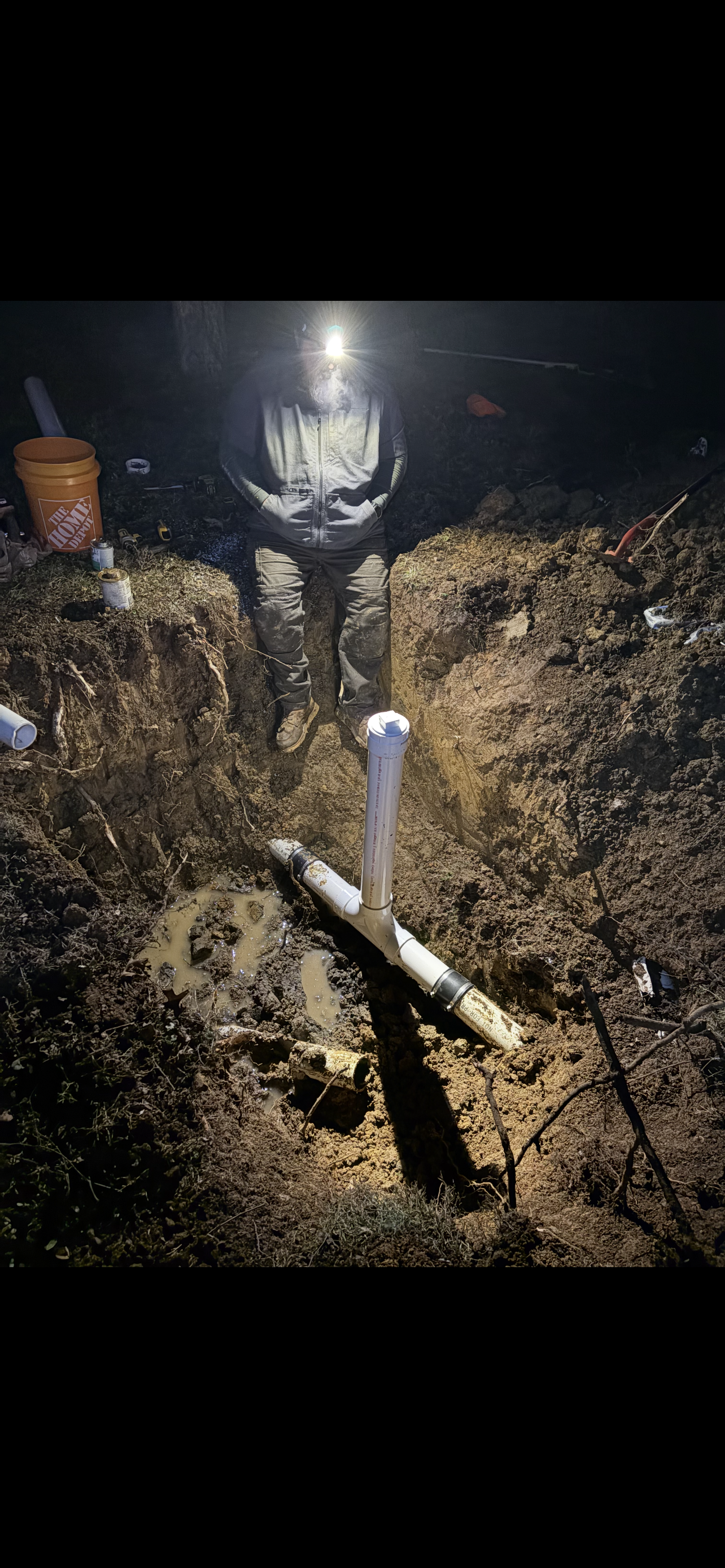 A person in a trench working on PVC pipes, lit by a headlamp. Dirt surrounds the pipes and person.