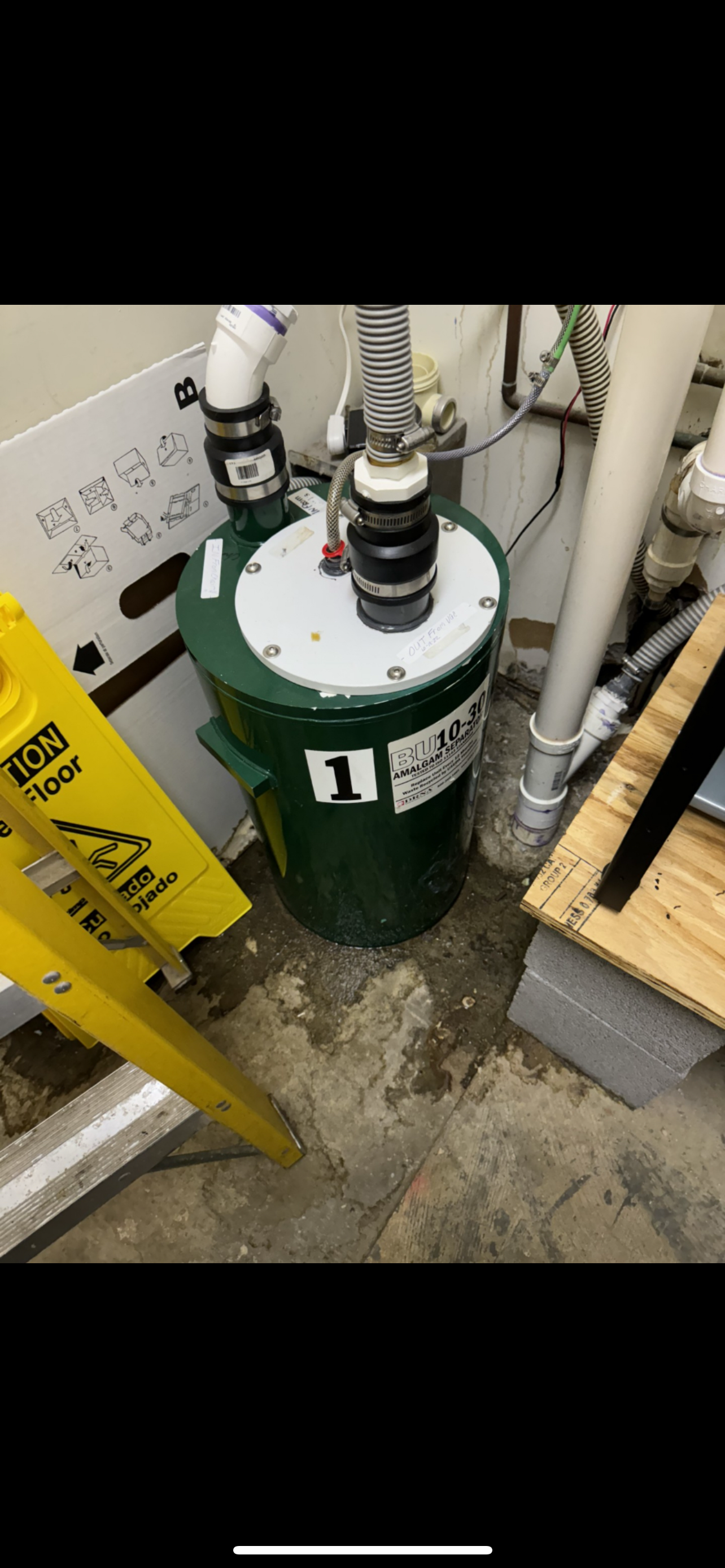 Green tank with pipes and wiring, in a concrete basement. Yellow caution sign leaning nearby.