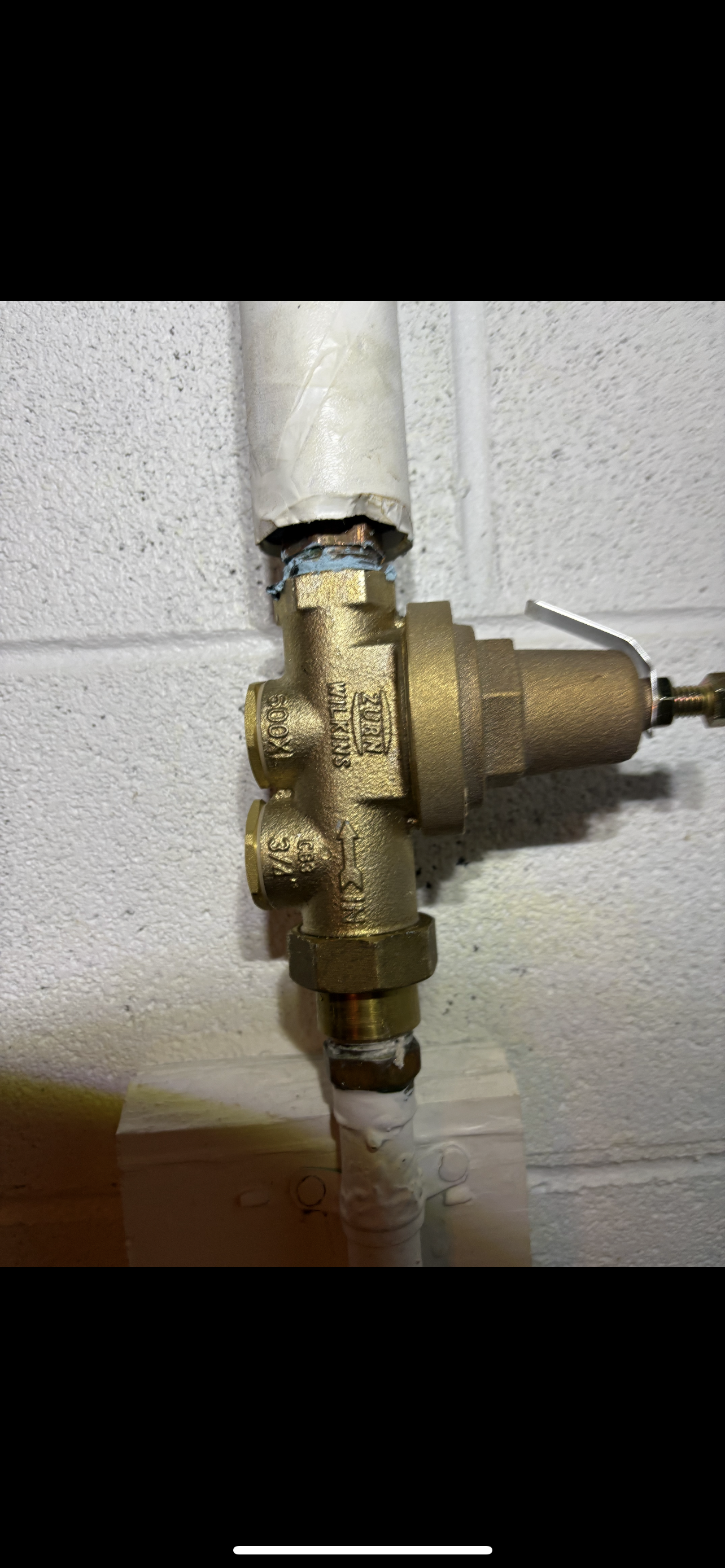 A brass water pressure regulator installed on a white pipe attached to a brick wall.