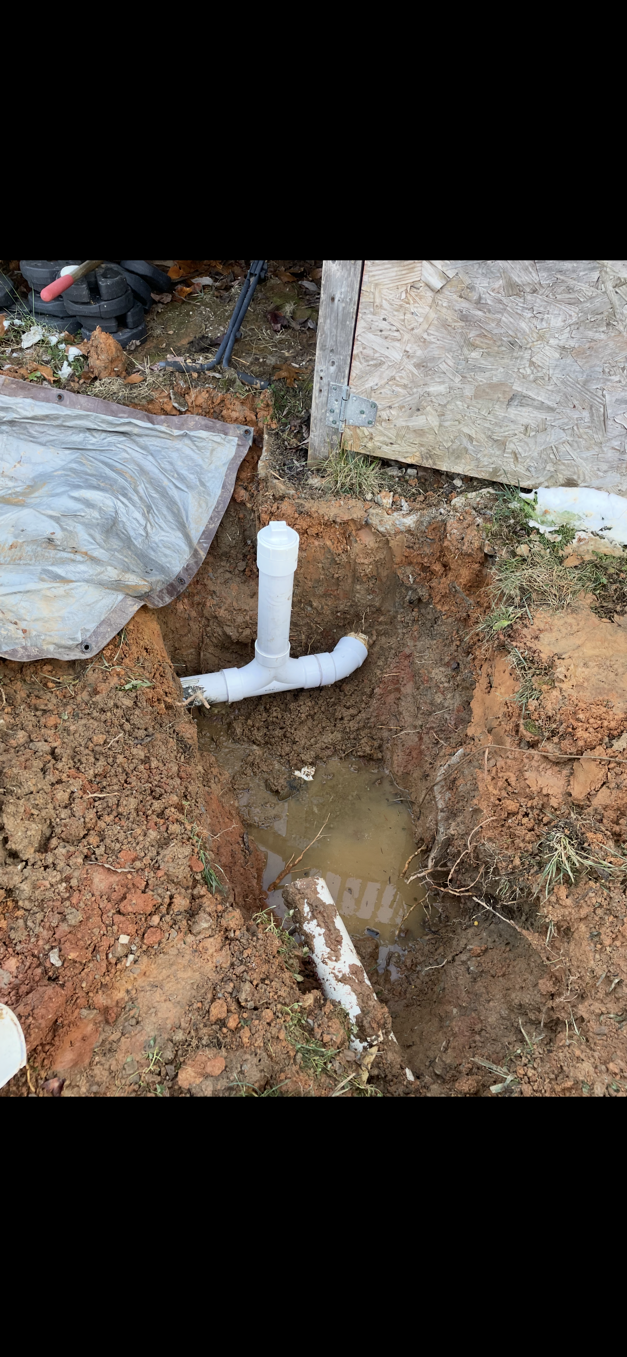 A T-shaped white pipe sits in a dirt trench with standing water. An open area, possibly an outdoor setting.
