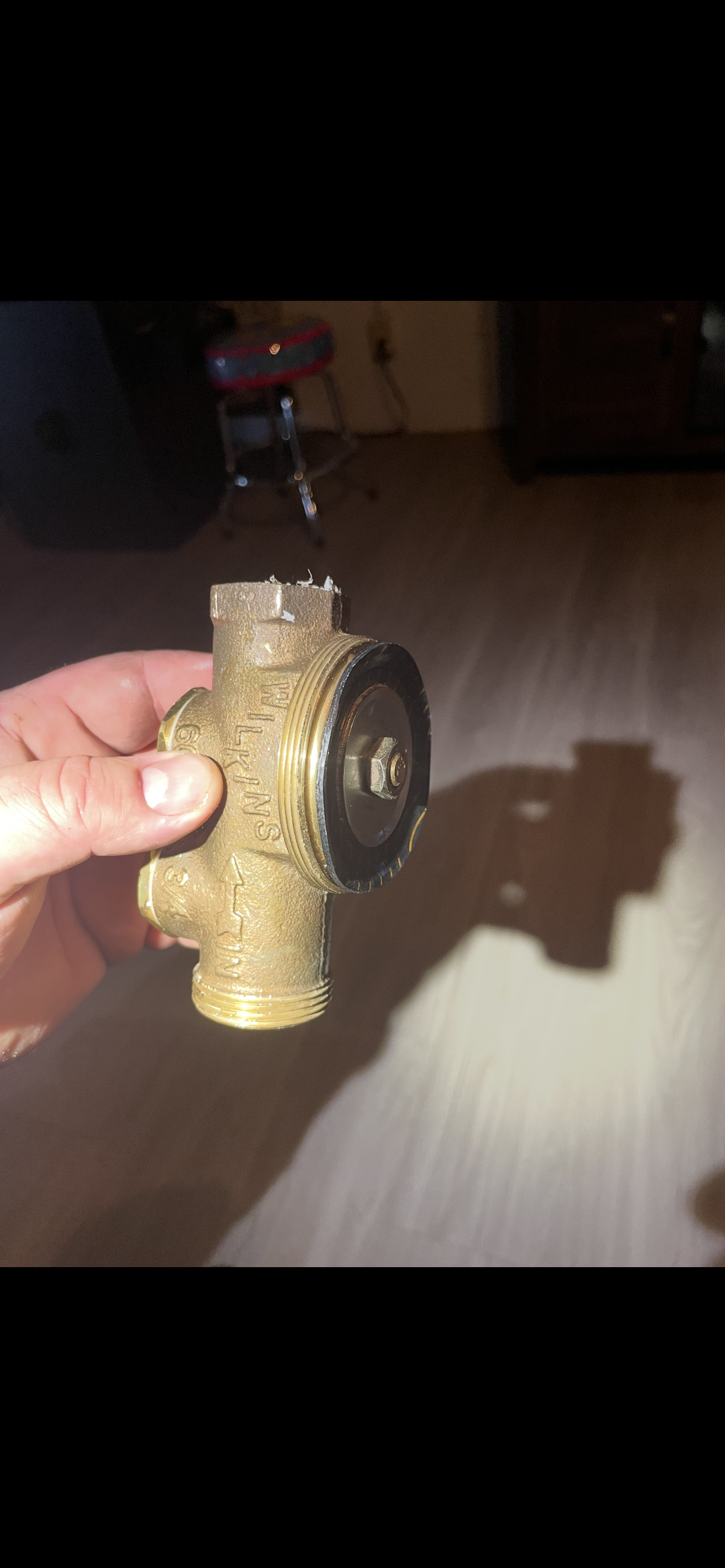 A hand holds a brass valve with a black rubber seal. A shadow is cast on a wooden floor.