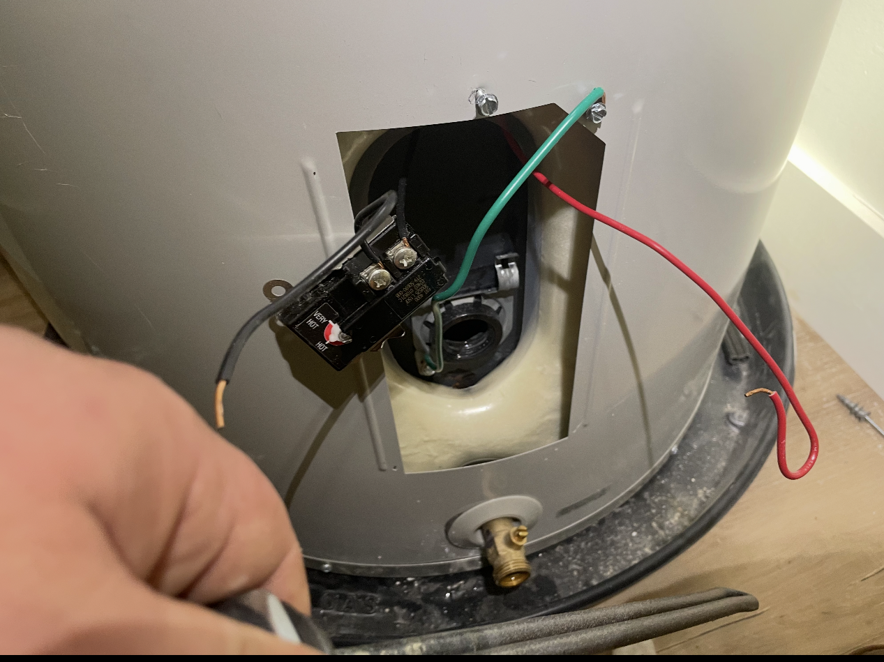 A person working on a water heater. The top access panel is open, showing electrical components and wiring.