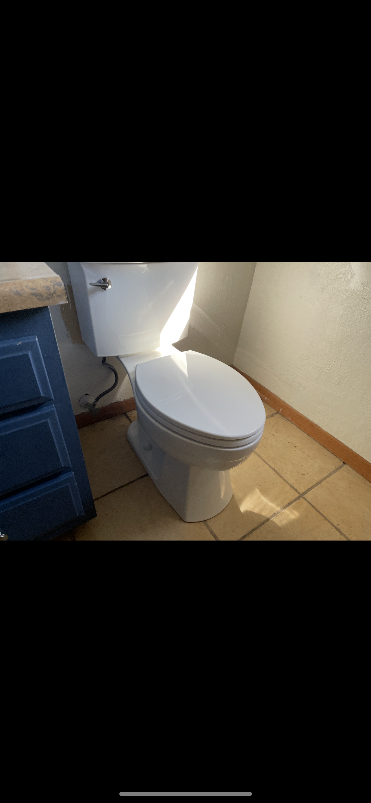 A toilet in a bathroom with blue cabinets and tan tile flooring, illuminated by sunlight.
