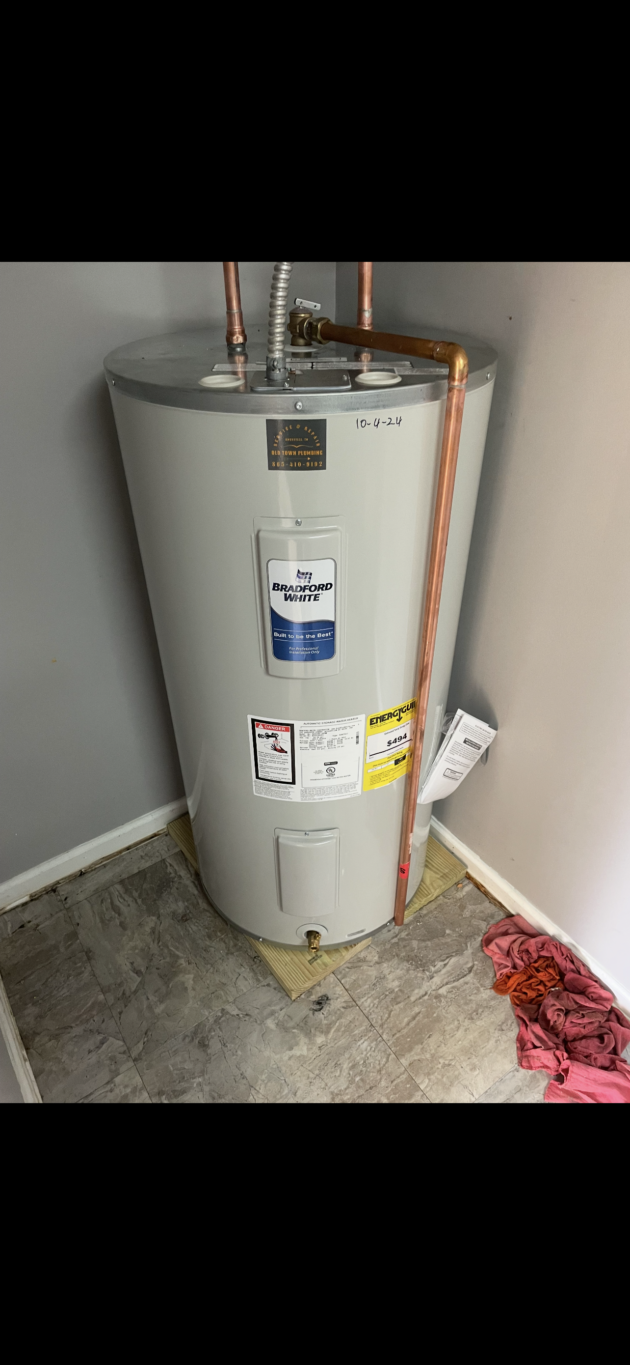 A tall, cylindrical water heater in a corner against gray walls. Copper pipes are visible. The floor is concrete.
