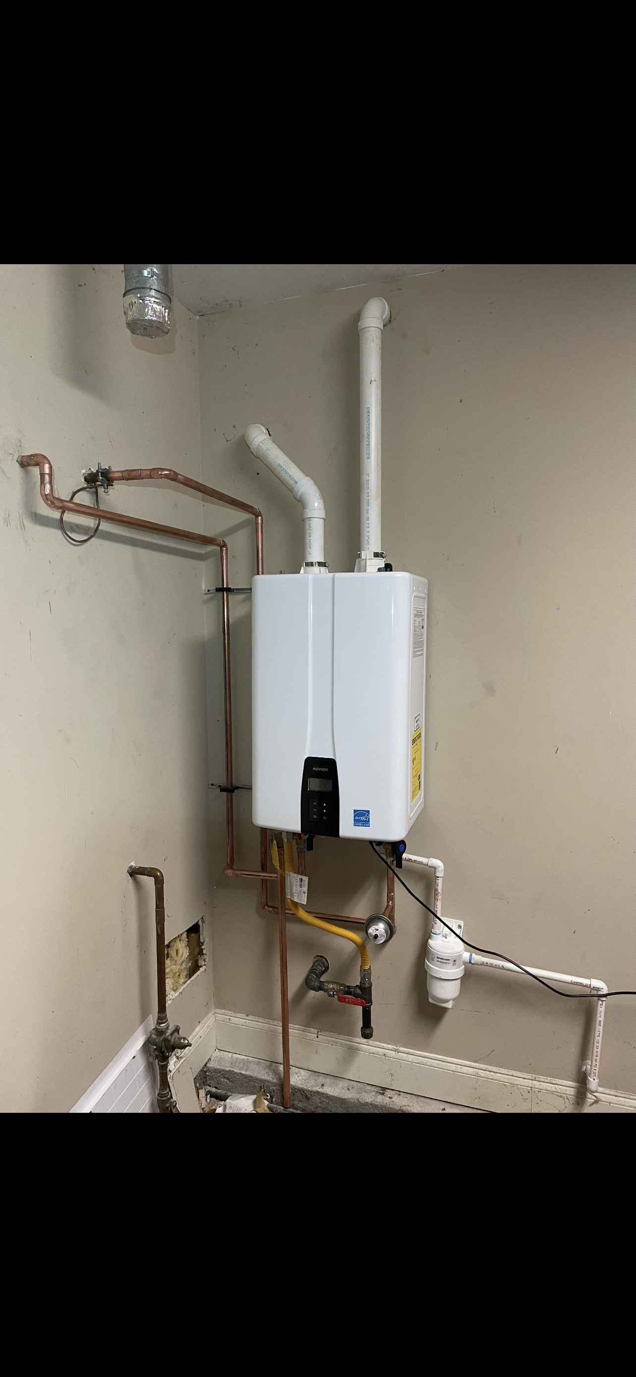 A tankless water heater installed on a wall, with copper and PVC piping.