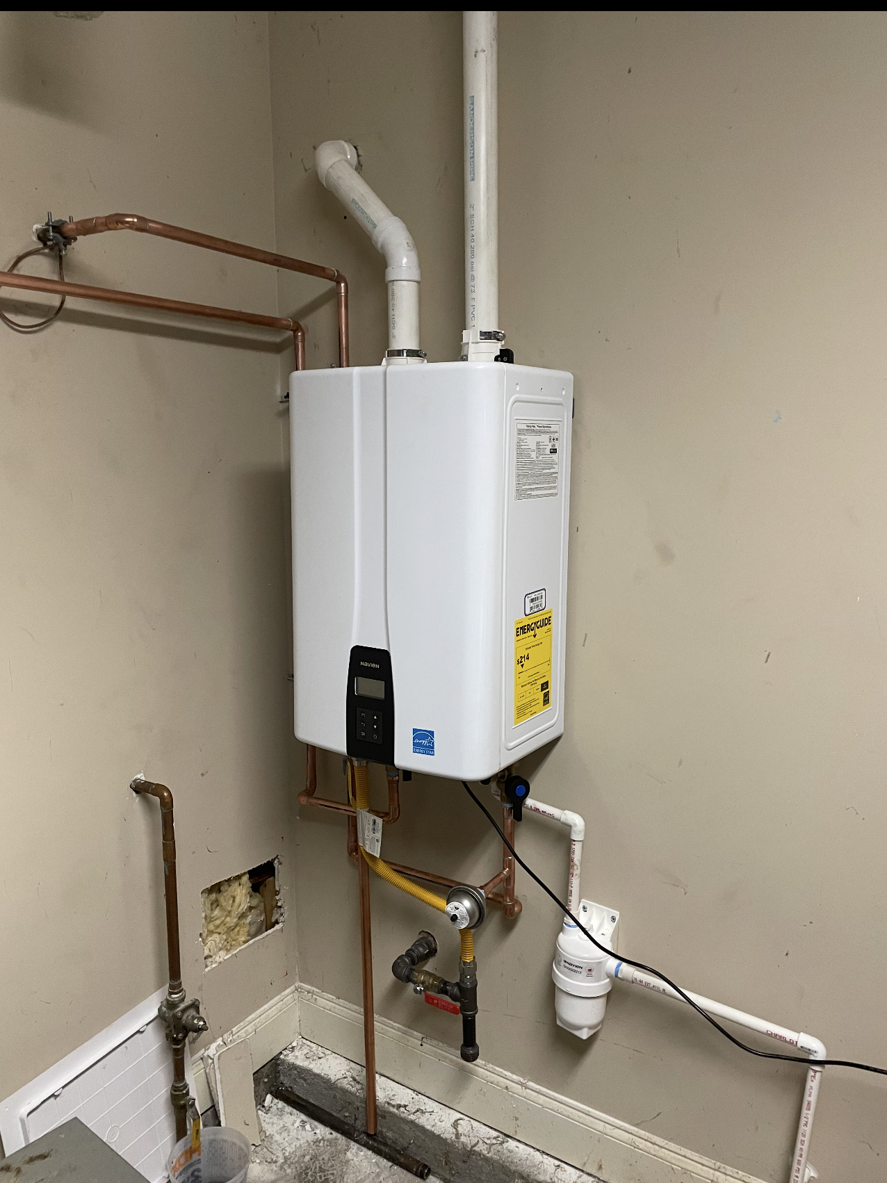 White wall-mounted tankless water heater with copper and PVC pipes in a utility area.
