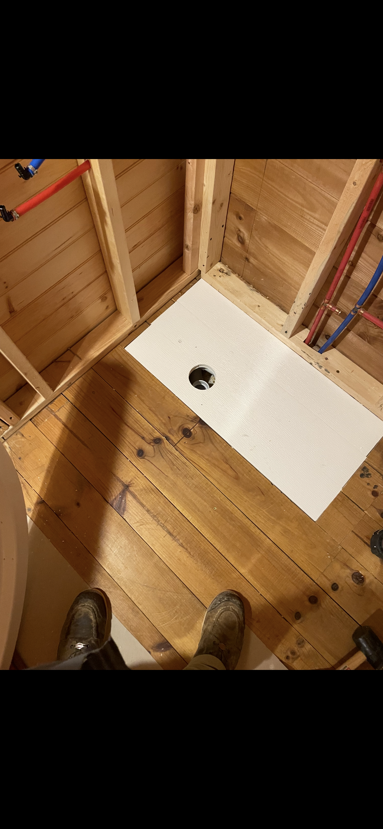 Wooden bathroom under construction with a white shower pan and exposed pipes.