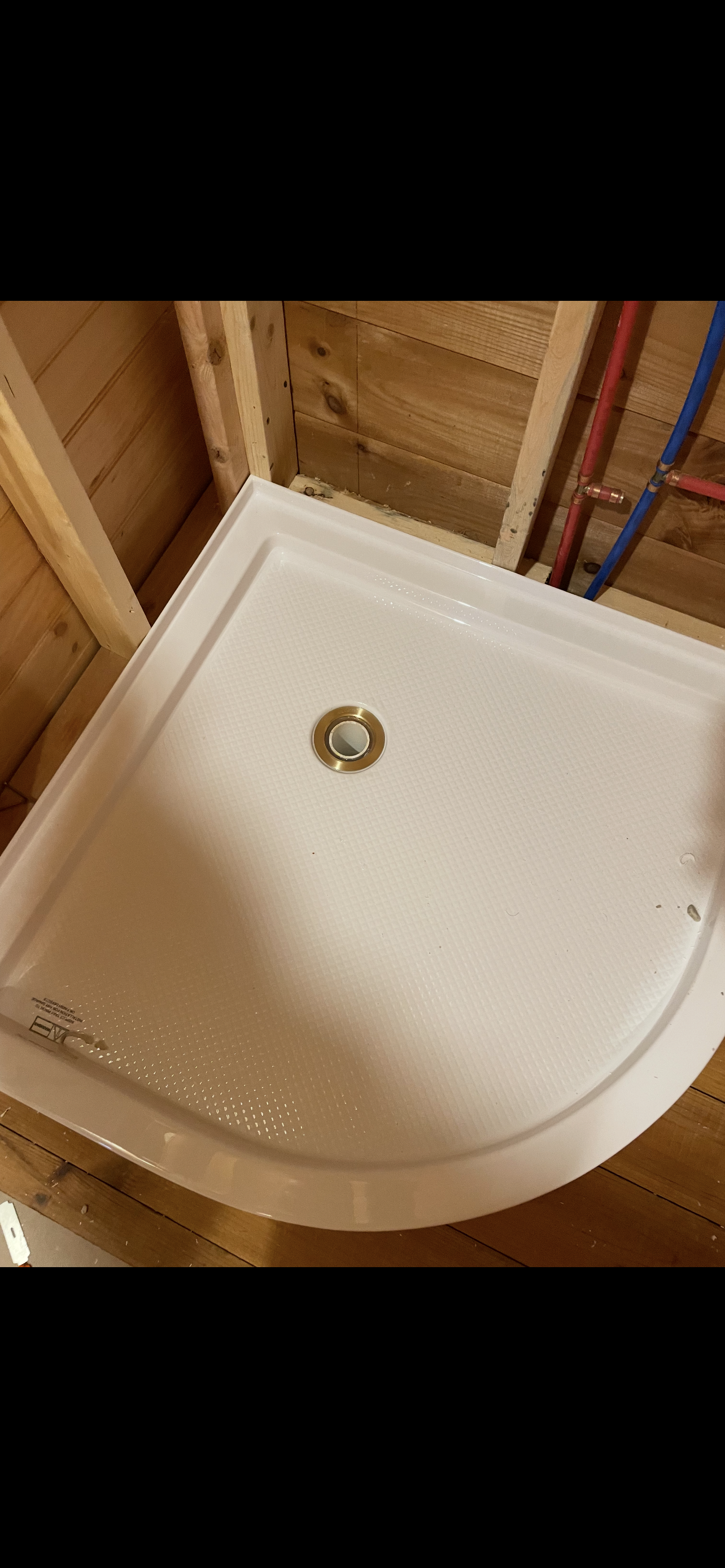 A white corner shower base installed, framed by wood beams in a basement, with drain and supply pipes visible.