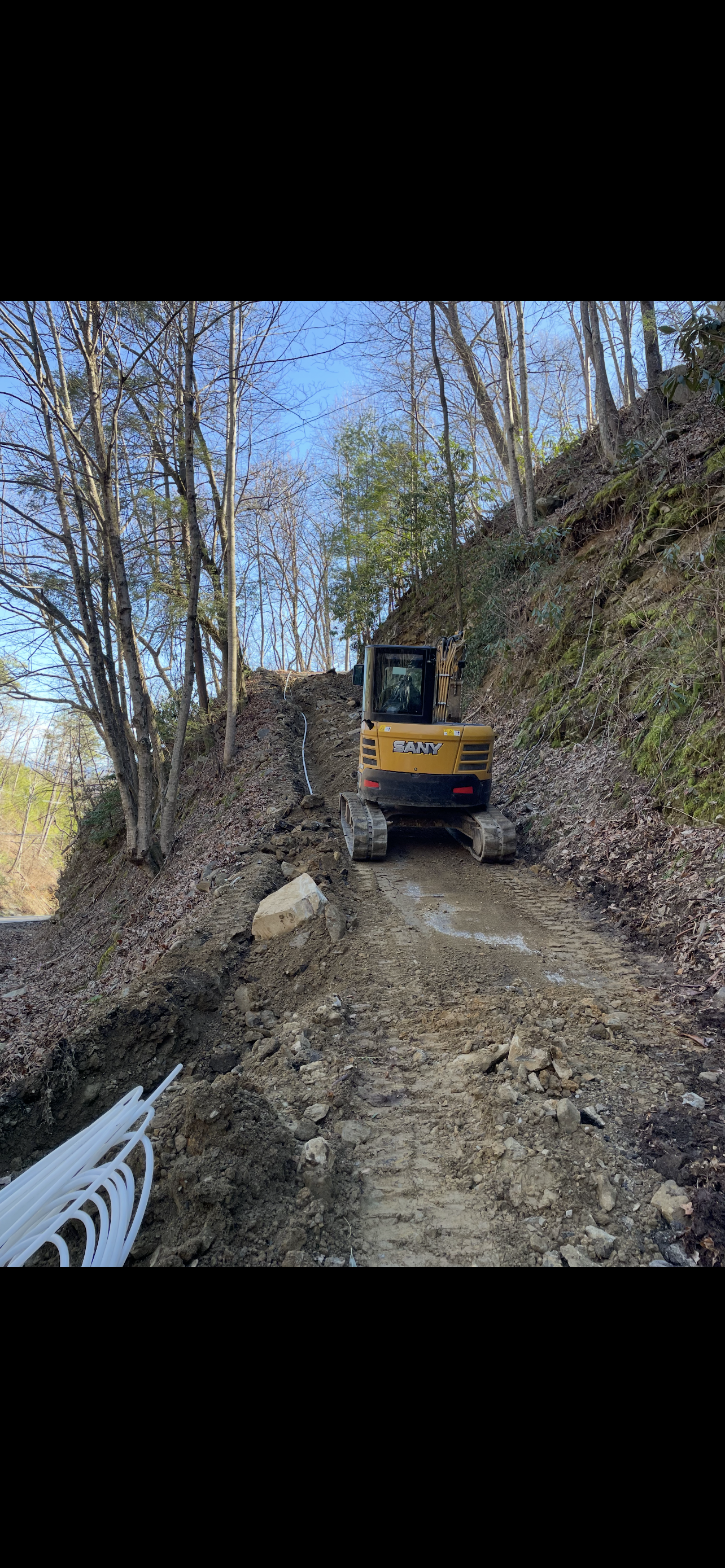 An excavator on a dirt road, clearing debris on a hillside, trees surround.