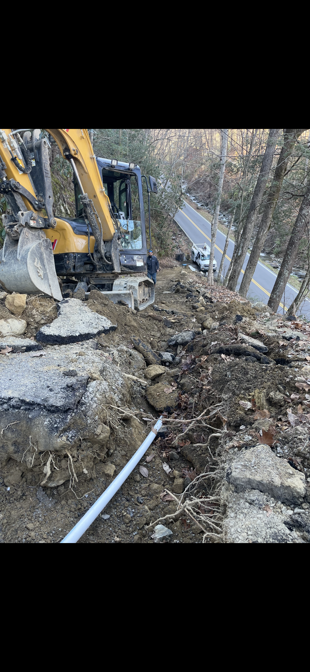 An excavator working on a road project.  Debris, trees, and asphalt are visible.