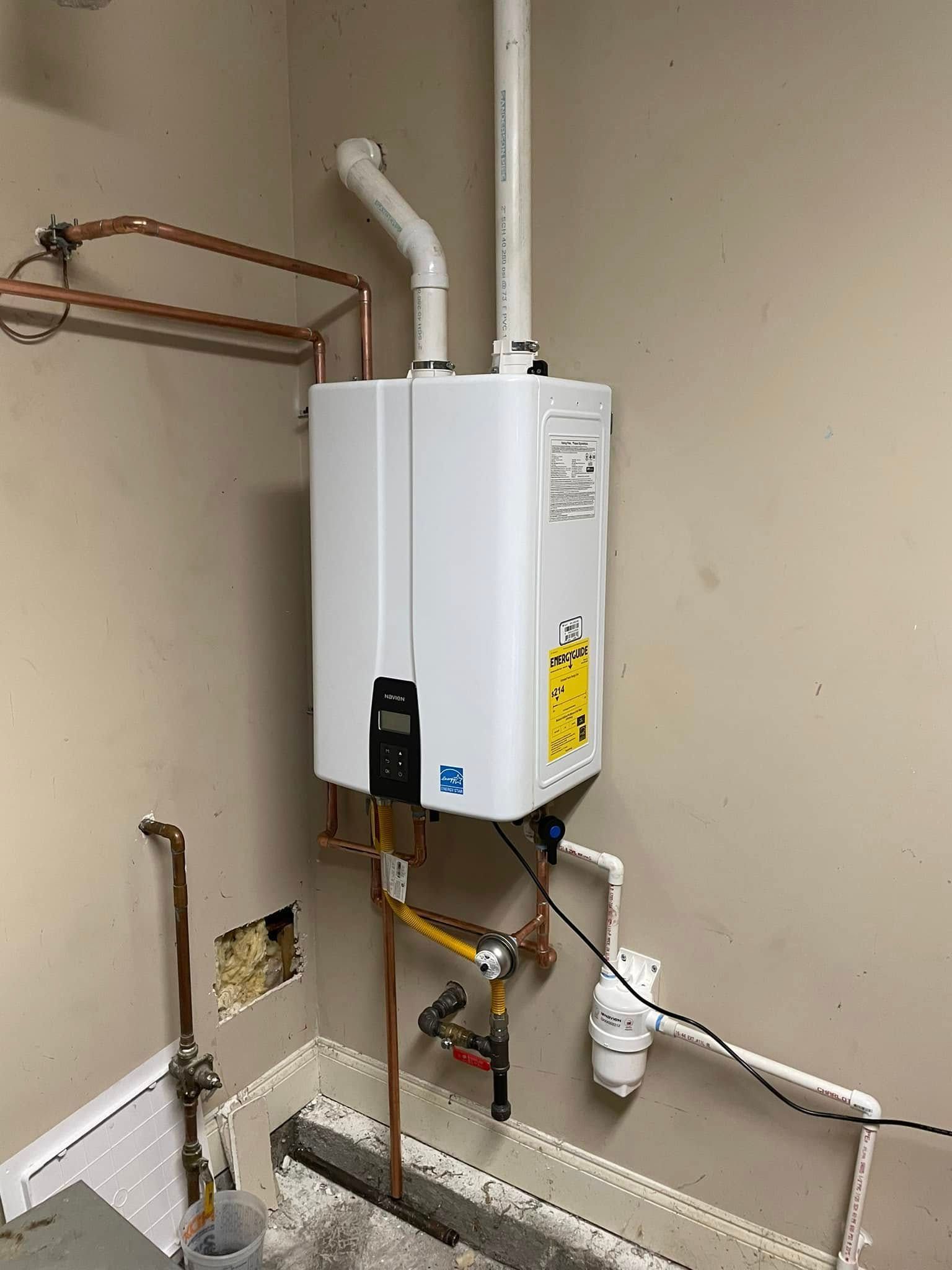 A white tankless water heater mounted on a beige wall with connected copper and white PVC pipes and a power cord.