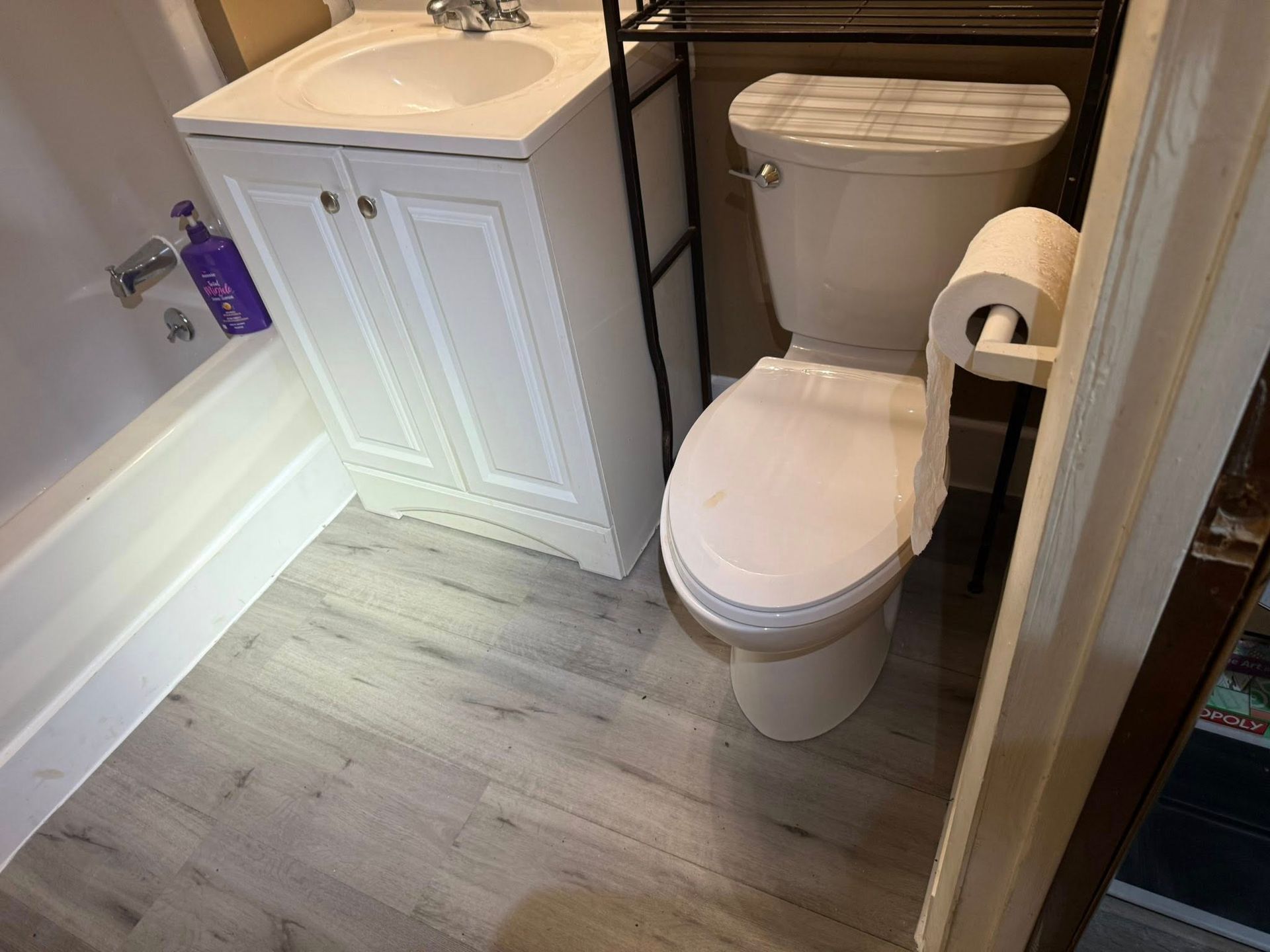 Small bathroom with toilet, sink, and bathtub. Light gray flooring, white fixtures, and a black shelf above the toilet.