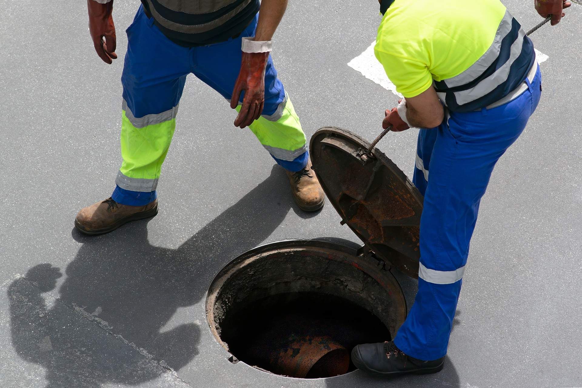 Two workers in high-visibility uniforms open a circular manhole cover on an asphalt road.